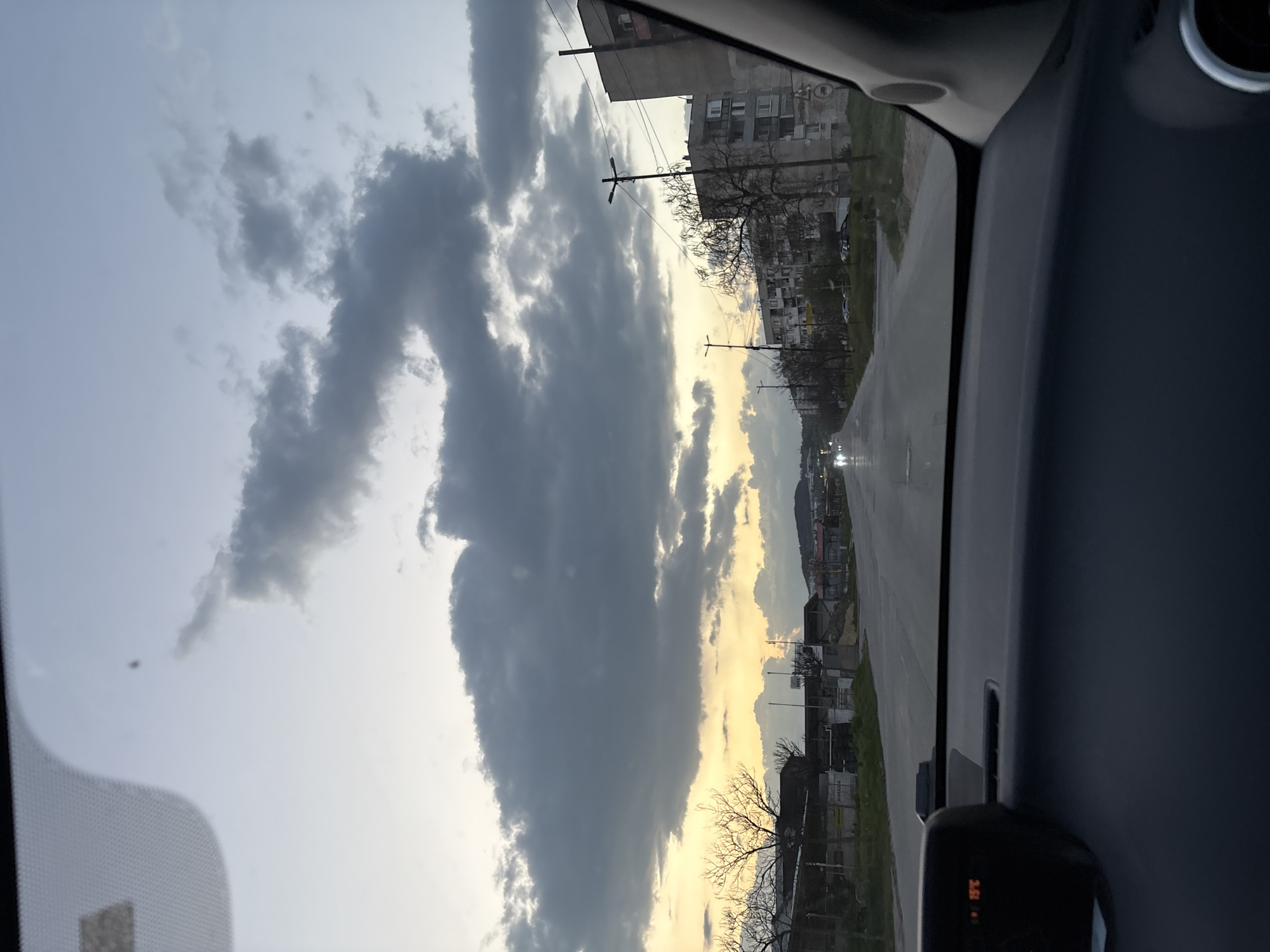 The image shows a view from inside a car driving on a wet road in Panagyurishte, Bulgaria. The sky is filled with heavy, dark clouds, although there is also a band of lighter, sunset-like colors visible on the horizon. Buildings line the roadside, with trees, some without leaves, interspersed between them. Power lines can be seen running along the street. The dashboard and part of the car's interior are visible.