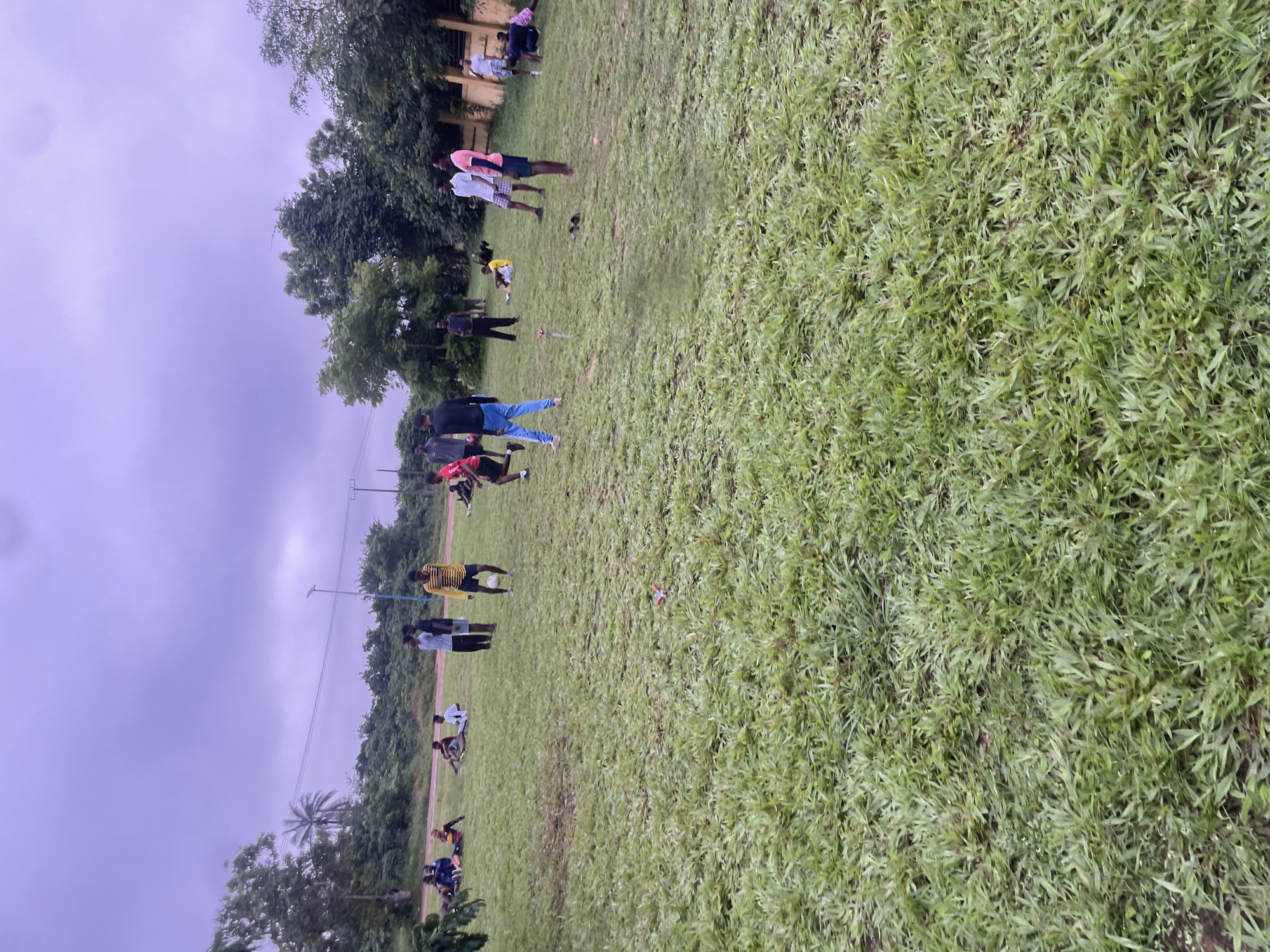 In Ihiagwa, Nigeria, a group of young men are gathered on a grassy field. The sky is overcast, with a purplish hue. In the foreground, the grass is lush and green, with some bare patches visible. A soccer ball is present on the field, suggesting a game or practice is underway. Several individuals are standing, while others are seated or lying down on the grass. One person is in the process of kicking the soccer ball. In the background, trees and foliage line the edge of the field, and a building with a yellow exterior is visible. There are also power lines and a lamppost in the distance. The scene appears to be a casual gathering of young men engaging in outdoor activity.
