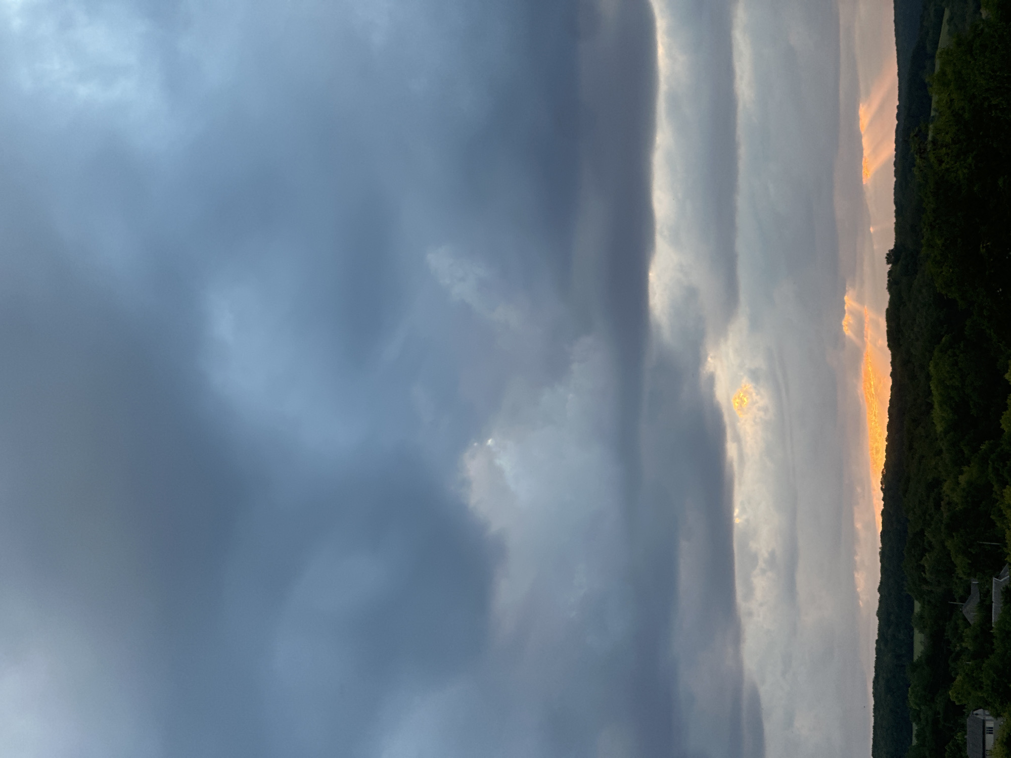 This is a photograph of a cloudy sky over a rural landscape. The sky is filled with dark, heavy clouds, with a band of orange light breaking through near the horizon, casting rays of sunlight down onto the land. Below the sky is a dense band of dark green trees that form a horizon line. In the foreground, there are a few small buildings, possibly houses, partially visible amongst the trees on the left side of the image. The overall impression is one of a dramatic, perhaps stormy, sunset or sunrise in a rural area. The location is stated to be Makiv, Ukraine.