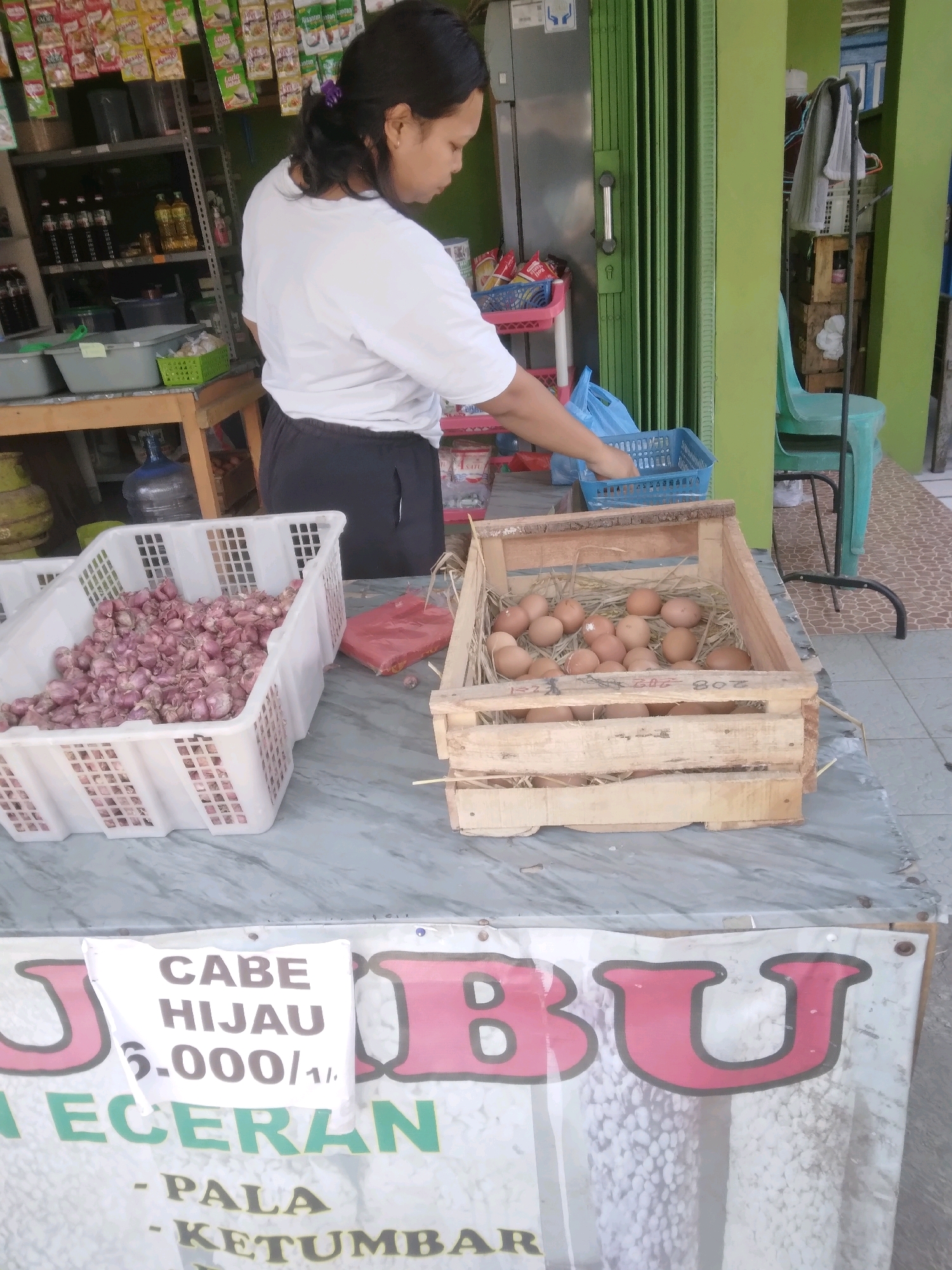 In Desa Pamijen, Kecamatan Sokaraja, Indonesia, a woman stands behind a table displaying various produce and food items. She is wearing a white t-shirt and dark pants and is in the process of arranging items in a blue plastic basket. On the table, a large white plastic crate is filled with red onions, and next to it, a wooden crate holds a collection of brown eggs nestled in straw. A white sign attached to a banner in the foreground displays text, including "CABE HIJAU" and pricing information. The background shows shelves stocked with goods, a green folding door, and a turquoise chair. The scene appears to be an outdoor market stall.