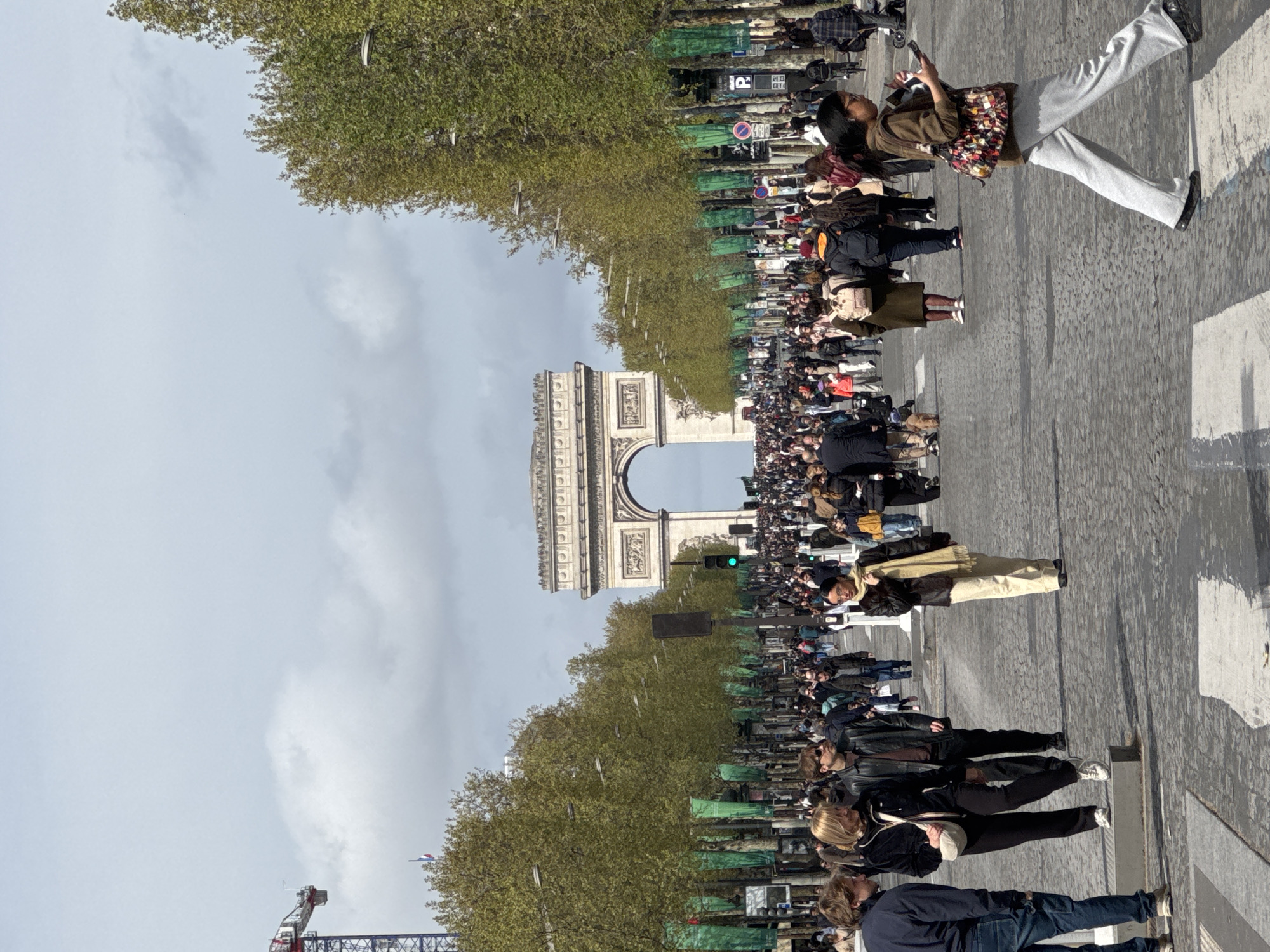 The image shows the Arc de Triomphe in Paris, France, viewed from the Champs-Élysées. A large crowd of people fills the avenue, some walking and some standing. Trees line both sides of the street, and streetlights are visible. A construction crane is visible in the lower left corner of the image. A traffic light is on.