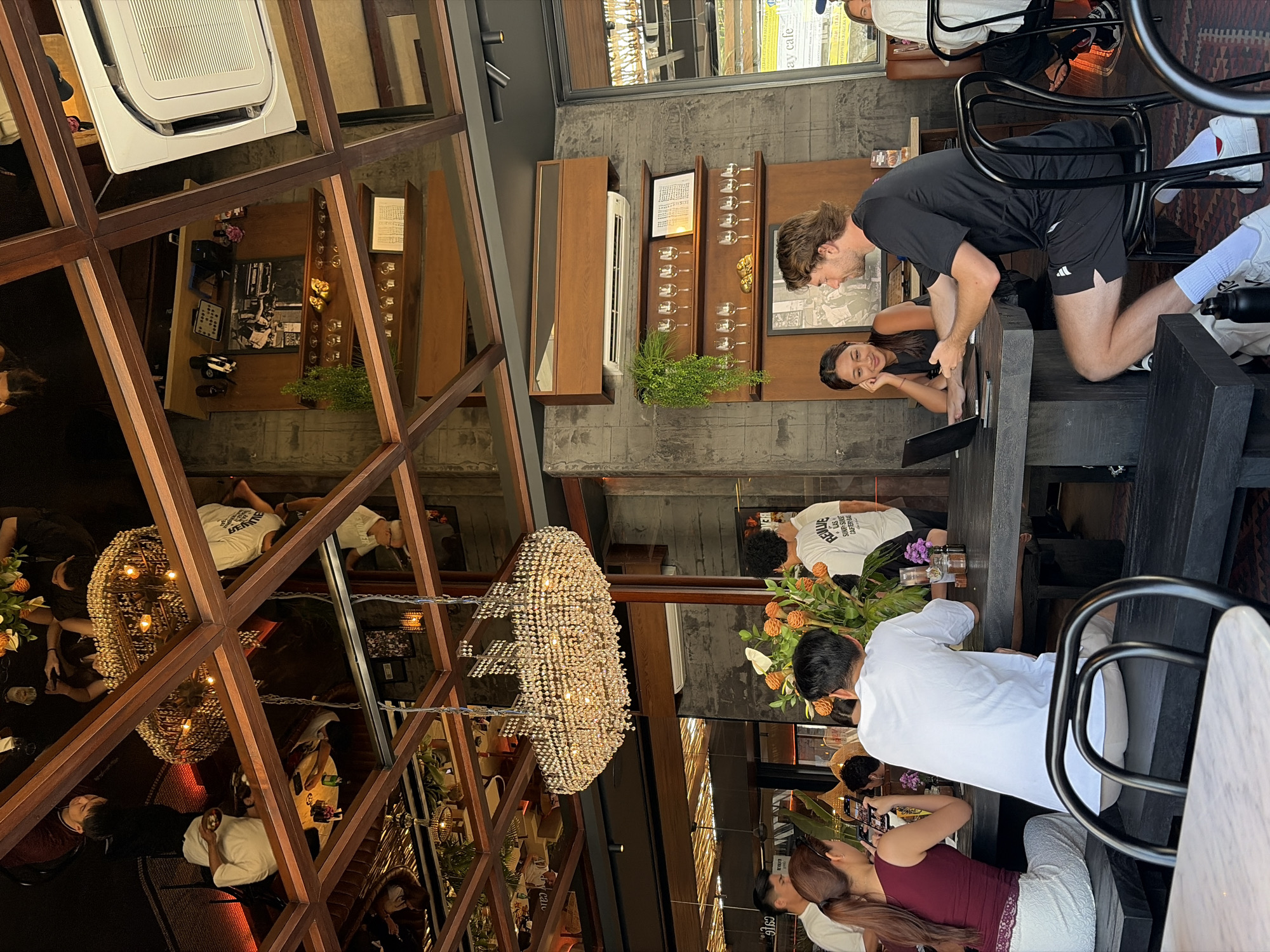 The image depicts the interior of a contemporary cafe in North Kuta, Indonesia. A large, ornate crystal chandelier hangs from a mirrored ceiling, reflecting the wooden beam structure above. Several patrons are seated at dark wooden tables. In the foreground, a woman in a maroon top and white skirt is looking at her phone. Next to her, a man in a white t-shirt faces away from the camera. To the right, a woman in a black tank top and a man in a black t-shirt and shorts are seated at another table, with a laptop open between them. The man is gesturing while speaking. The cafe features concrete walls, wooden shelves displaying glassware, and plants. An air conditioning unit is visible mounted high on the wall. The overall atmosphere suggests a relaxed, modern dining environment.