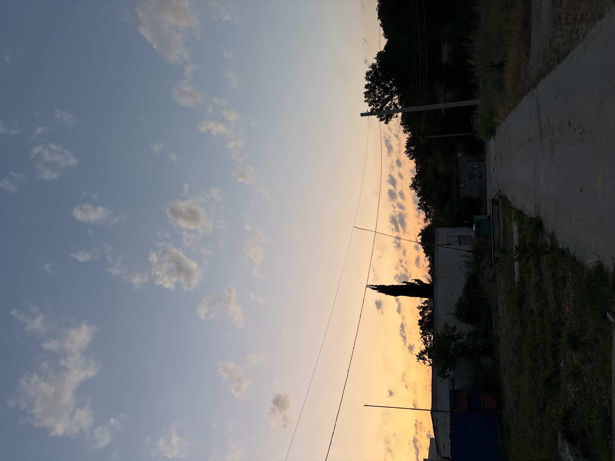 An outdoor, low-angle, vertical shot depicts a rural scene at dusk in Malyi Mayak, Ukraine. The sky is a gradient of pale blue at the top transitioning to a soft orange glow near the horizon, dotted with scattered white clouds. A solitary, dark, tall, slender tree stands silhouetted against the sky, just to the left of the image's vertical center. Below the tree, a long, low, white building with a dark roof occupies the middle ground. To the left of the building, a tall, dark blue rectangular structure and a smaller, dark red rectangular structure are visible. A concrete path leads from the foreground into the distance, bordered by dry grass and vegetation. In the background, a dense line of dark green trees forms a natural border. A single utility pole with visible wires stands to the right of the path, extending upwards into the sky. The overall lighting suggests late evening or early morning twilight.