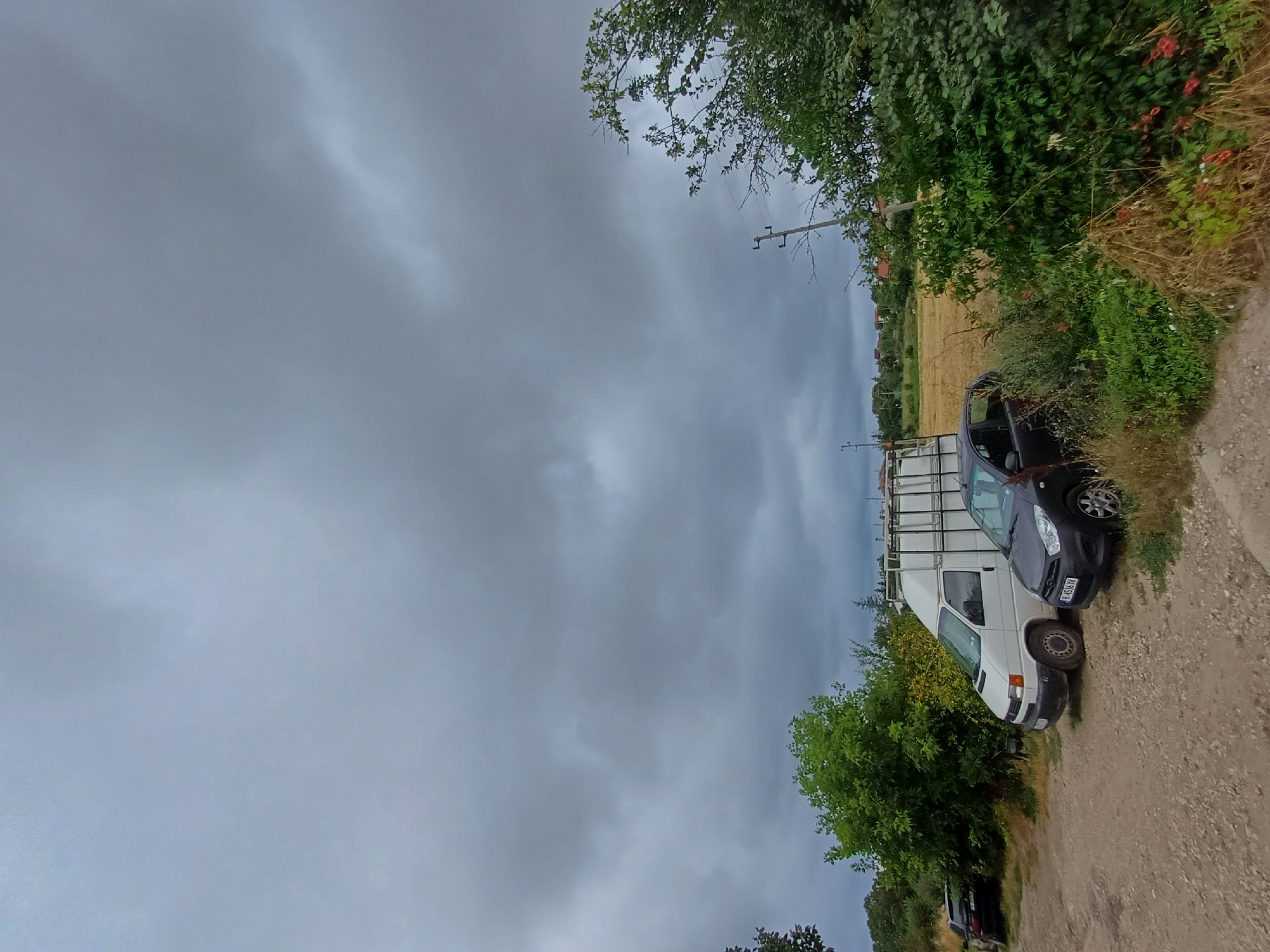 A low-angle, eye-level shot captures a cloudy day with dark, ominous clouds filling most of the sky. In the foreground, a dirt road leads to a parking area where two vehicles are parked. To the left, a white panel van is parked next to a dark-colored hatchback. The van appears to have a metal cage-like structure on top. Further to the left, a dark car is parked. Behind the vehicles, a line of green trees and bushes forms a dense backdrop, with a field of dry grass and crops visible in the distance. The overall scene suggests a rural or semi-rural setting, possibly on the outskirts of Varna, Bulgaria, given the style of the vehicles and the landscape. There are no people visible in the image. The environment is natural, with vegetation and a dirt road, indicating an outdoor, undeveloped area.