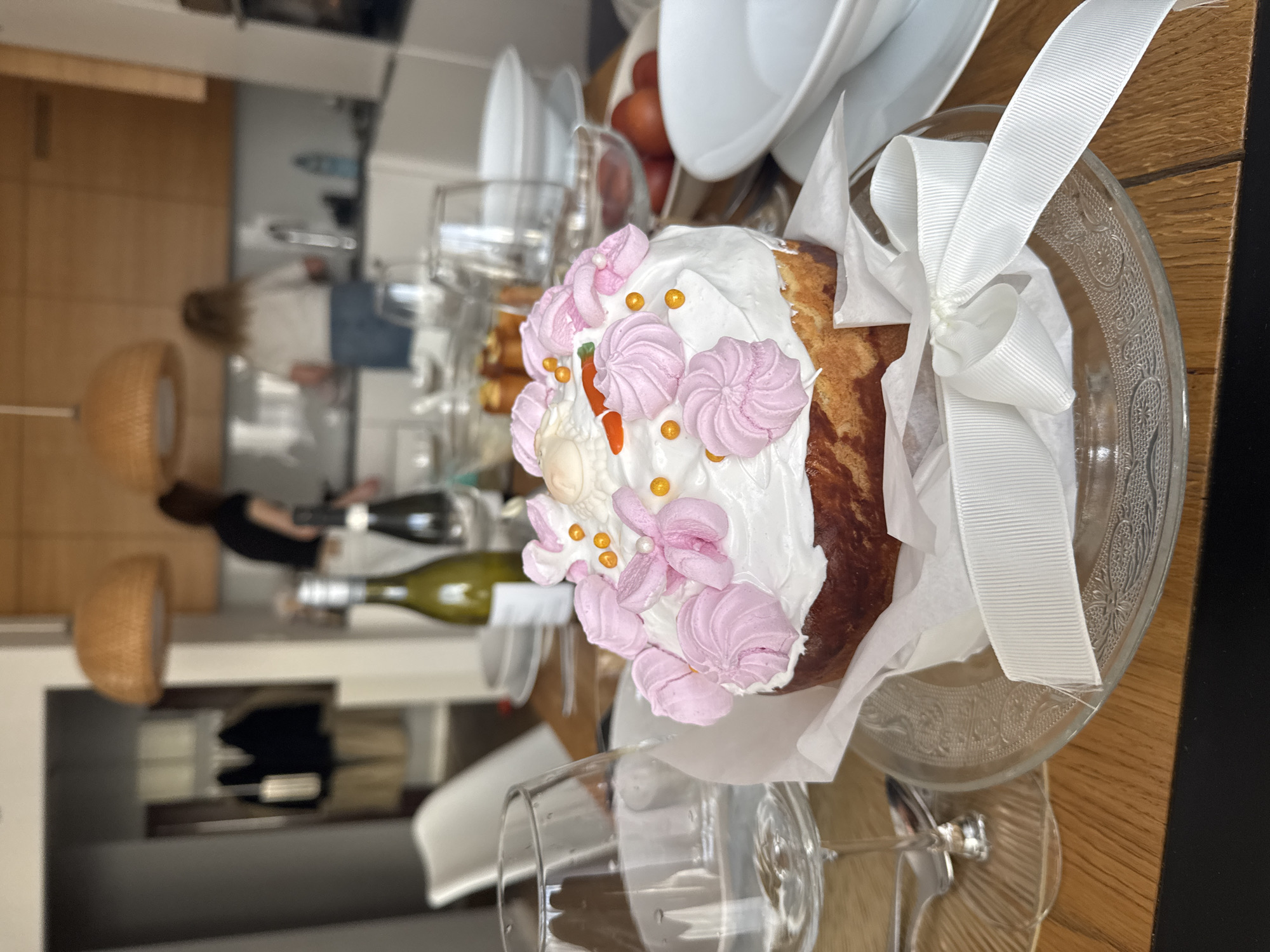 Here is a description of the image:
A slightly tilted close-up shot focuses on an elaborately decorated Easter cake (Paska) set on a clear glass serving plate. The cake is adorned with white frosting, pink meringue-like decorations, and small orange sprinkles, with a white ribbon tied around it. The cake is in paper packaging. It sits on a wooden surface, and a partially visible wine glass rests on the surface.
In the blurred background, more glassware and white dishes can be seen, along with blurred bottles of wine. Further back, a person in a black shirt can be seen in a kitchen setting with white cabinets. The room is lit with natural and warm artificial light from lamps.
While the exact location within Warsaw cannot be determined solely from this image, it depicts a dining scene with Easter cake.