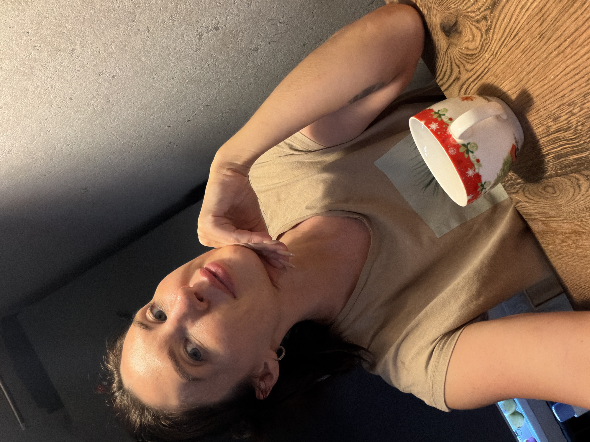A woman with brown hair and blue eyes, wearing a tan t-shirt, is sitting at a wooden table with a mug in front of her. She is resting her chin on her hand, looking towards the camera. The mug has a festive pattern with red and white colors. The background is a textured gray wall and a dark area. There is no indication of the city of Perm, Russia in the image.