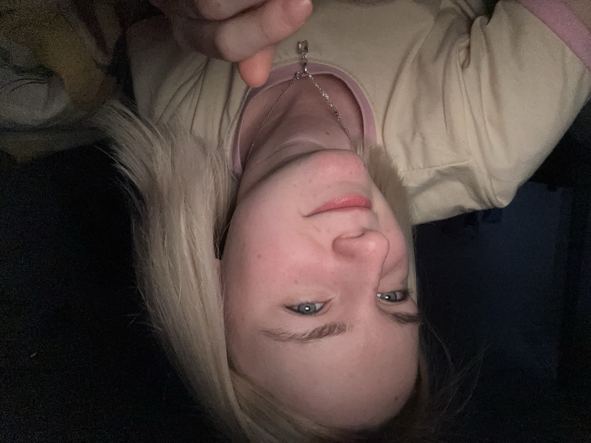 A close-up, slightly rotated image shows a woman with fair skin and light blonde hair. She has blue eyes and is wearing a light-colored shirt with pink trim around the neckline. A silver necklace with a small pendant is visible around her neck. The background is dark and out of focus. There is no indication of the location being Vsevolozhsk, Russia.