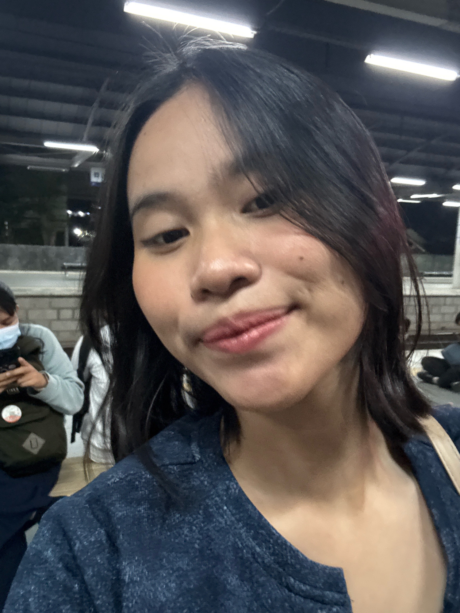 A woman is taking a selfie in an indoor public space, possibly a train station. She has dark hair and is wearing a dark blue jacket. In the background, another person wearing a mask and a backpack is visible, as well as a train platform and overhead lighting. The environment appears to be dimly lit. The exact location in East Jakarta, Indonesia cannot be determined from the visual information provided in the image.