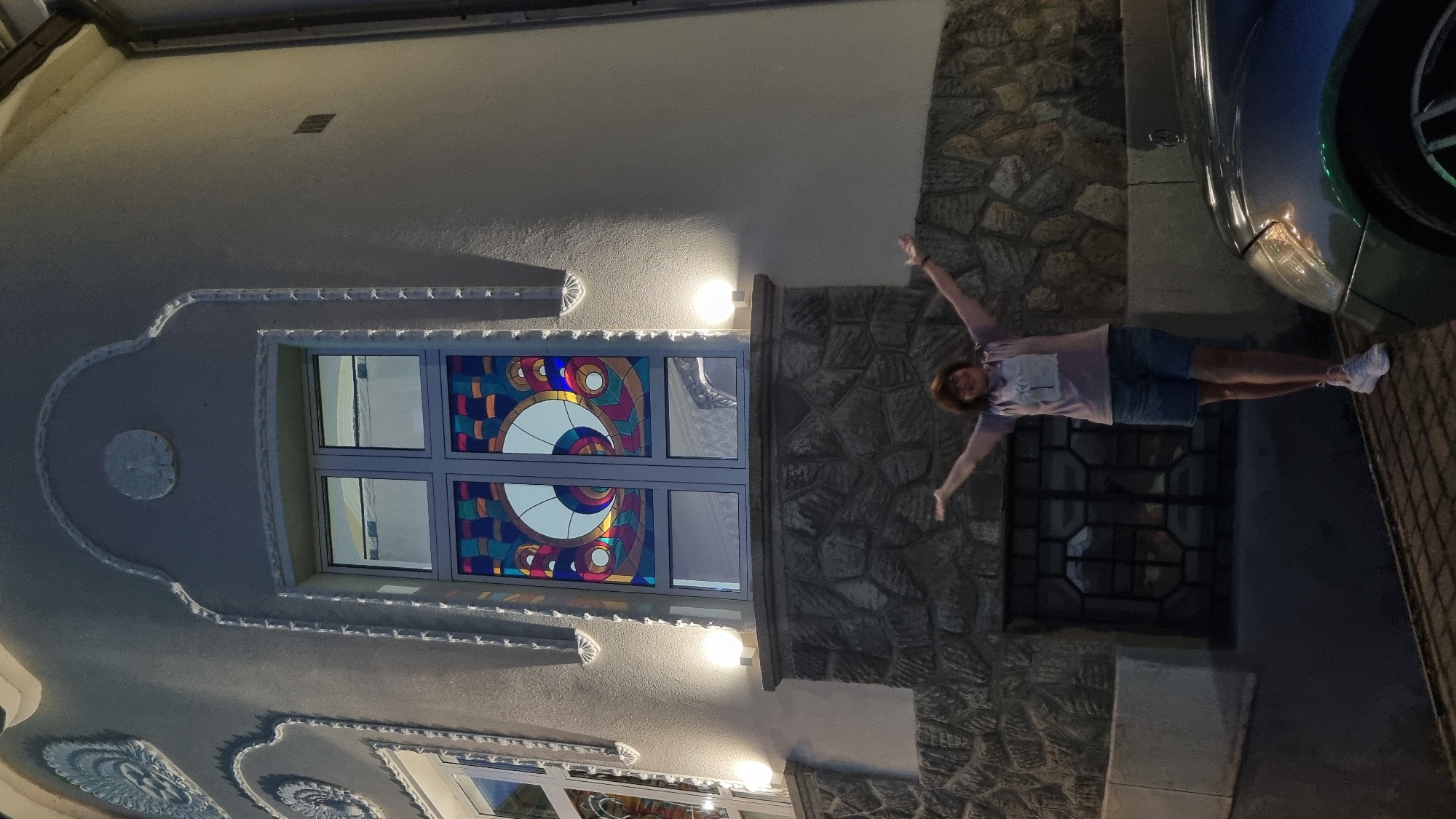 A woman stands with her arms outstretched in front of a building with a stained glass window and stone facade. The woman is wearing a purple t-shirt and denim shorts, and white sneakers. The building appears to be a residential or public building, with decorative molding and lighting. There is a car parked in the foreground, partially visible. The image was taken at night, as indicated by the artificial lighting and the dark sky. The architecture of the building, particularly the stained glass window and decorative elements, suggests it may be located in Berehove, Ukraine, a city known for its Art Nouveau architecture.
