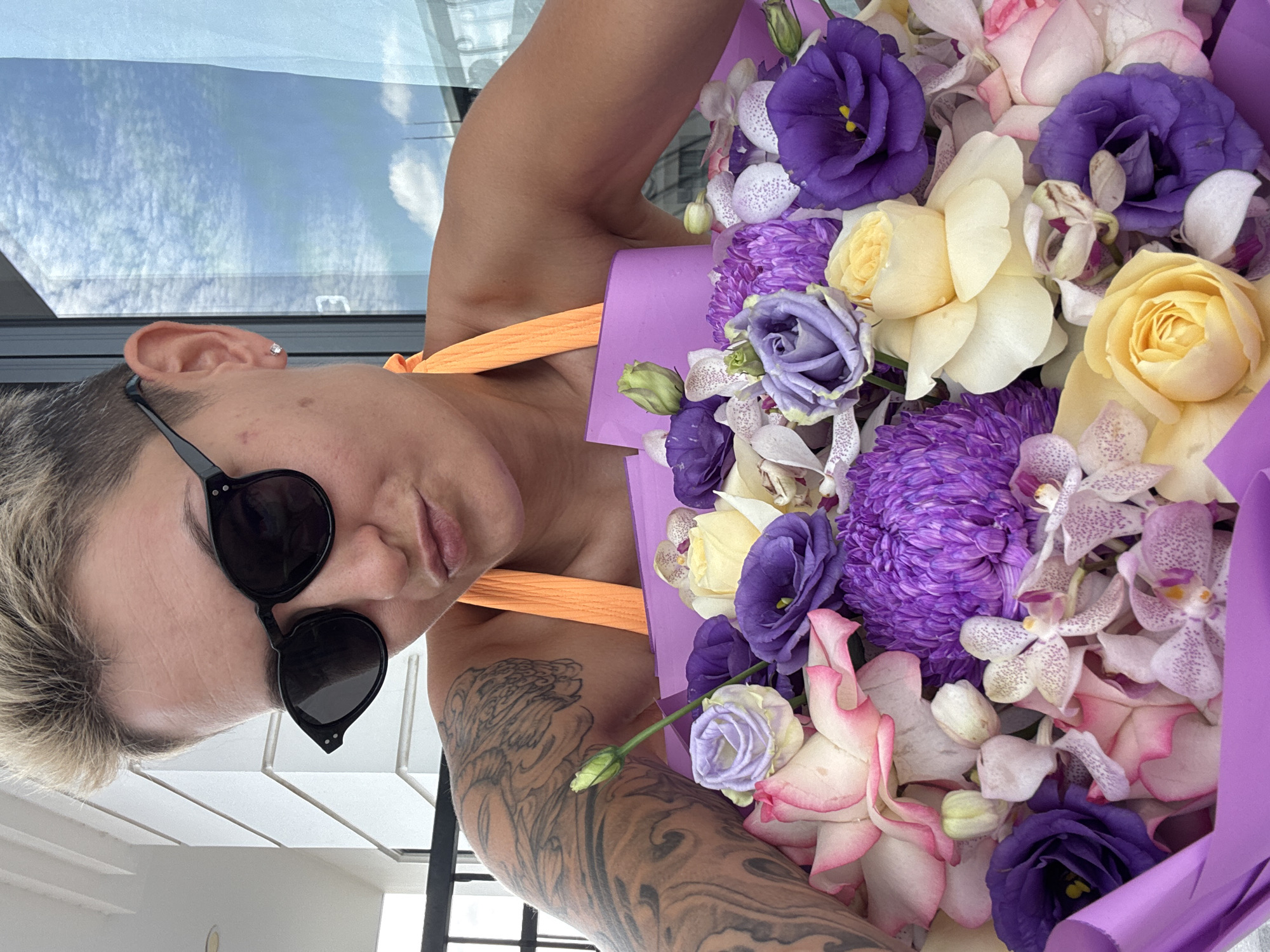 An individual with short, blonde, spiky hair, wearing black sunglasses and an orange halter top, poses for a selfie outdoors, holding a large bouquet of purple and pink flowers. The background reveals a balcony railing, a white building with architectural details, and a bright blue sky with scattered clouds. The person has tattoos on their left arm and is wearing a small, square earring in their left earlobe. The overall impression is a candid shot taken in a bright, sunny environment, possibly a hotel balcony or terrace in Nha Trang, Vietnam.