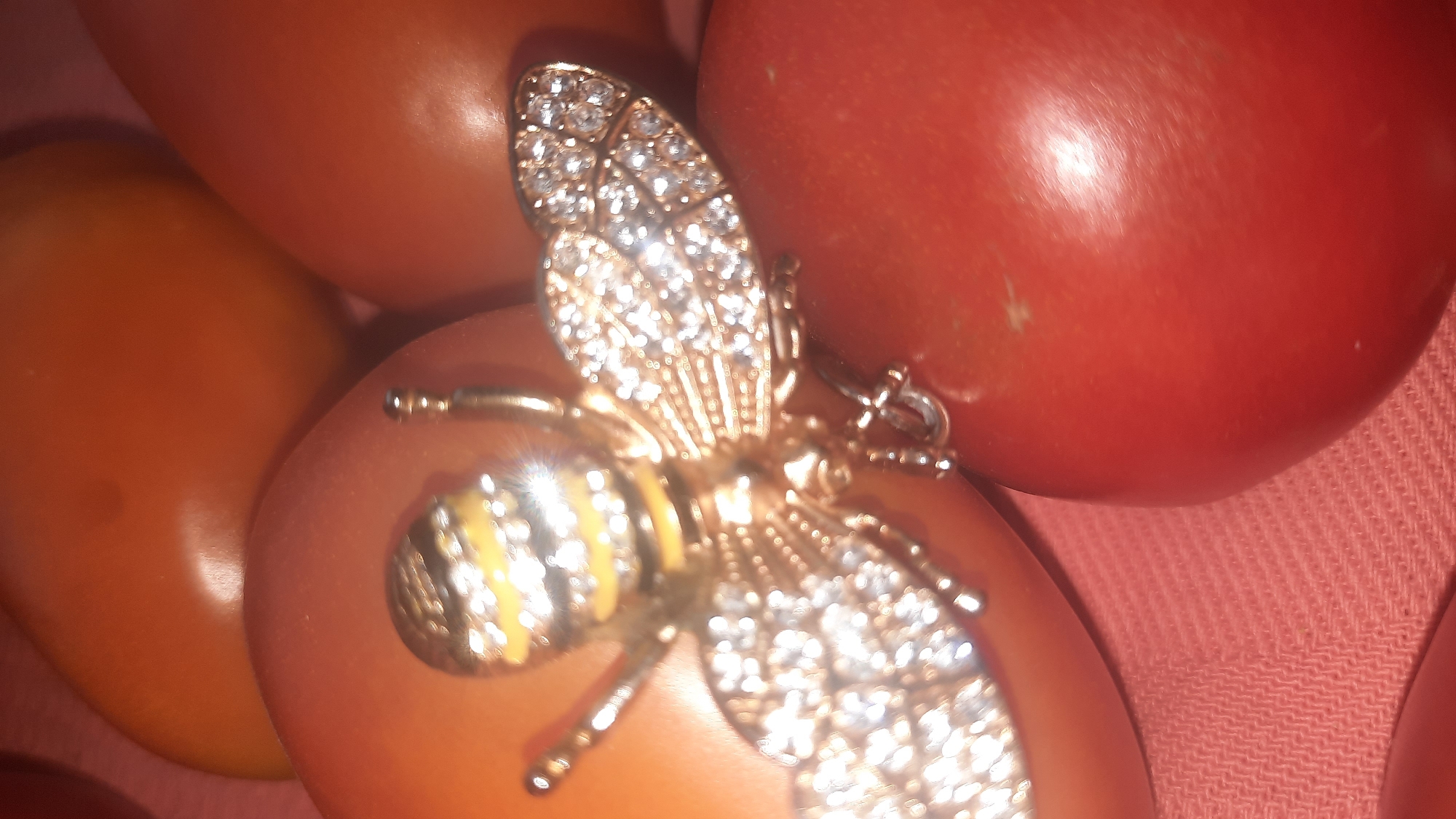 In a close-up, top-down view, a bejeweled bee brooch with gold accents and clear rhinestones rests on a cluster of tomatoes and a pink textured fabric. The brooch is positioned diagonally, with its head facing left and its wings spread. The tomatoes, some red and some orange-toned, are rounded and smooth, forming a base for the brooch. The fabric underneath appears to be a textured knit, possibly a tablecloth or placemat. The lighting creates highlights on the brooch and tomatoes, suggesting an indoor setting. There is no information to determine the location in the city of Шклоў, Belarus.