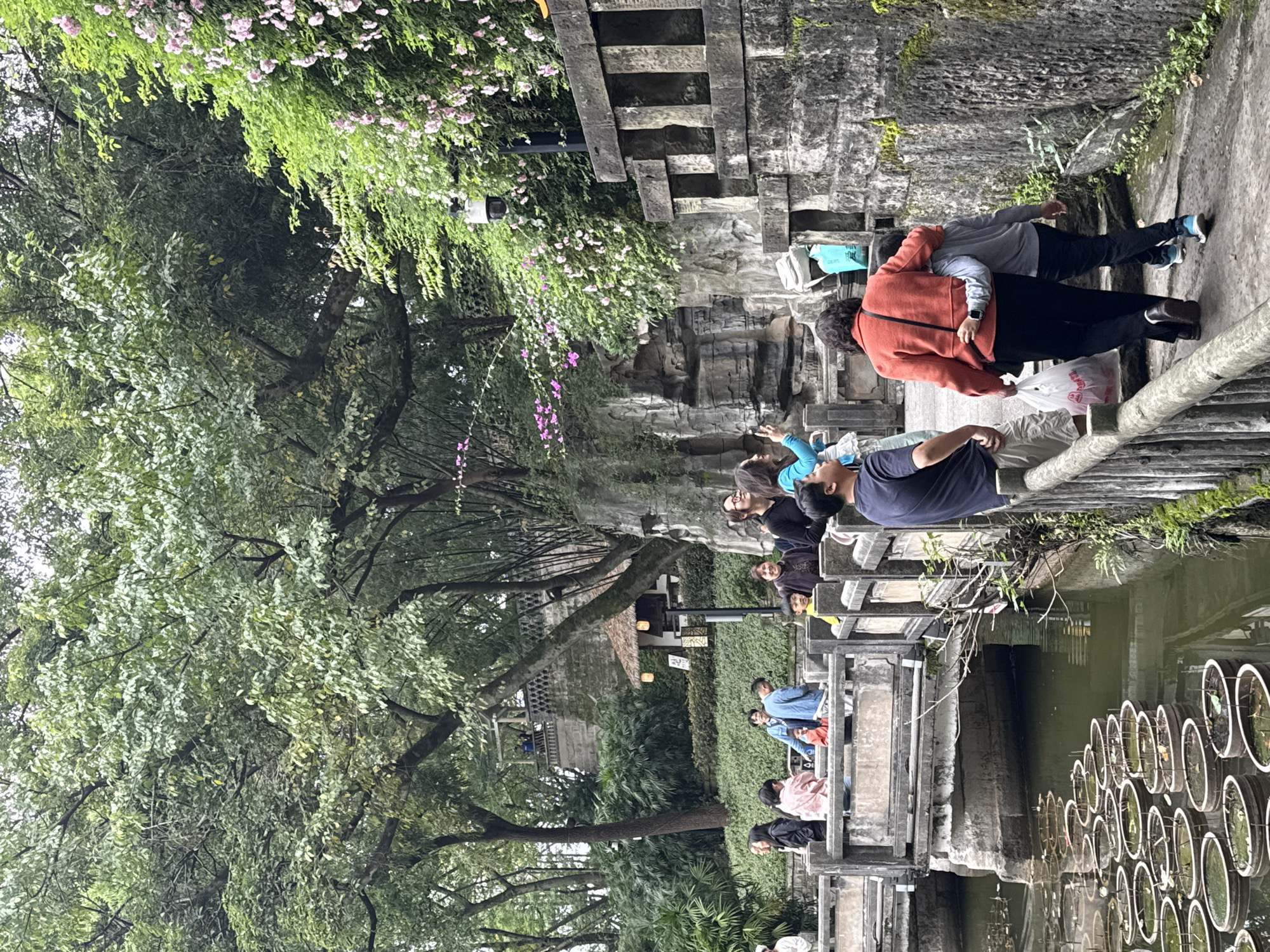 A horizontal shot captures a scene in the Yuzhong District of China. A group of people gather near a waterway on what appears to be a stone bridge. The bridge and surrounding structures are constructed of concrete and stone, with green flora, including trees and vines, visible on the left and above. In the waterway, numerous circular structures are partially submerged.
Several people are seen on the bridge. On the left, a few people are sitting, facing to the left, while others stand or move along the bridge. At least one person in a red top is visible. Two individuals are closely embracing near what may be a barrier.
The environment includes architectural elements such as stone steps, while a structure with a traditional roof is observed among the foliage. The lighting suggests an overcast day.