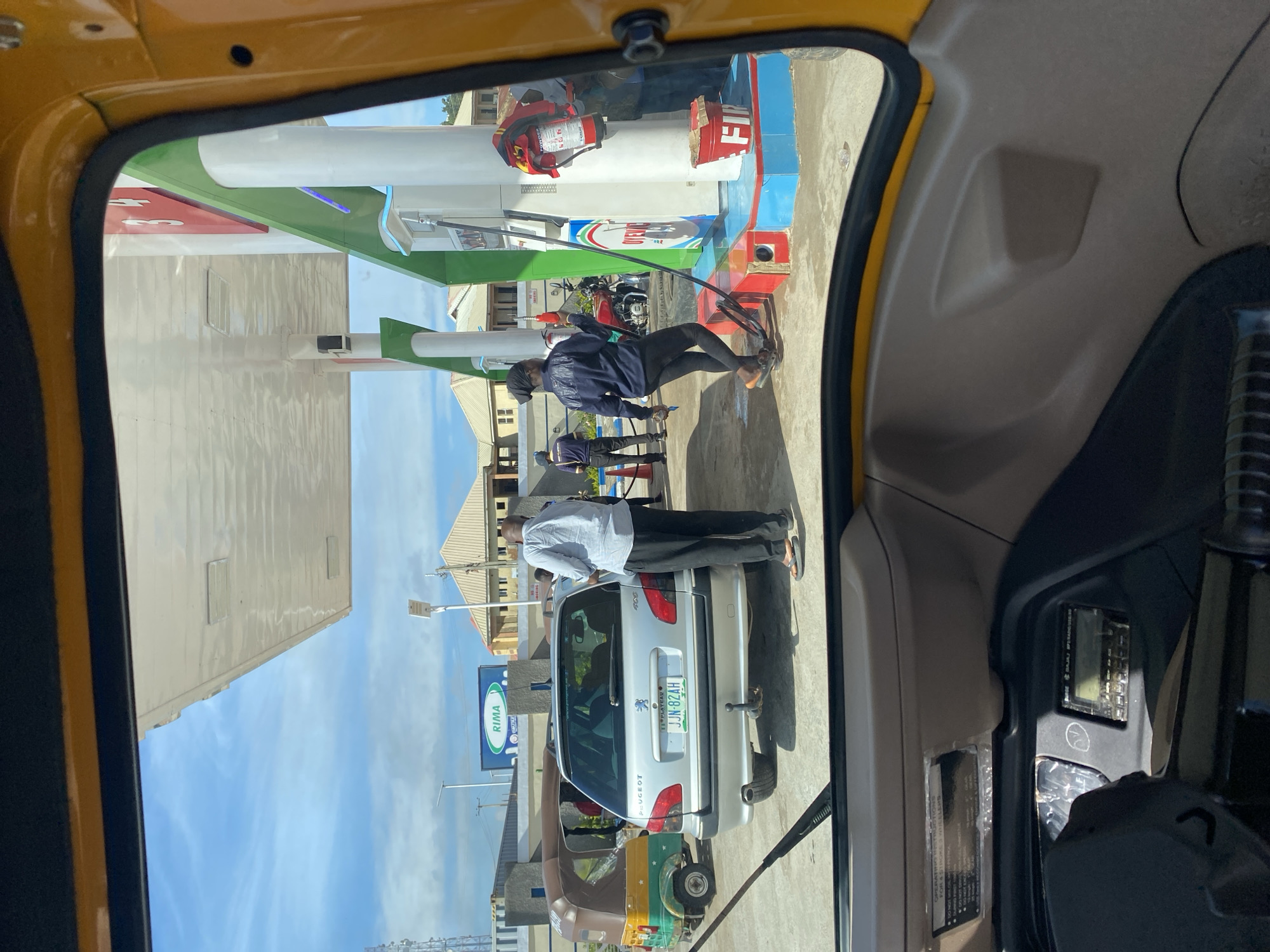 The image shows a scene at a gas station in Barakin Lerawi, Nigeria, viewed from inside a yellow auto-rickshaw. A silver Peugeot station wagon is parked at a gas pump, and a man in a light blue shirt and dark pants stands beside it, holding what appears to be a receipt. To the left of the Peugeot, a colorful auto-rickshaw is also present. In the background, a gas station canopy with white pillars and green accents is visible, along with buildings and a blue sky with clouds. Another auto-rickshaw, this one green and brown, is parked further to the left. A woman in a dark blue jacket and black leggings is operating a gas pump, holding a red nozzle. A fire extinguisher is mounted on one of the gas pump pillars. The interior of the auto-rickshaw from which the photo is taken is visible, including the dashboard and steering wheel.