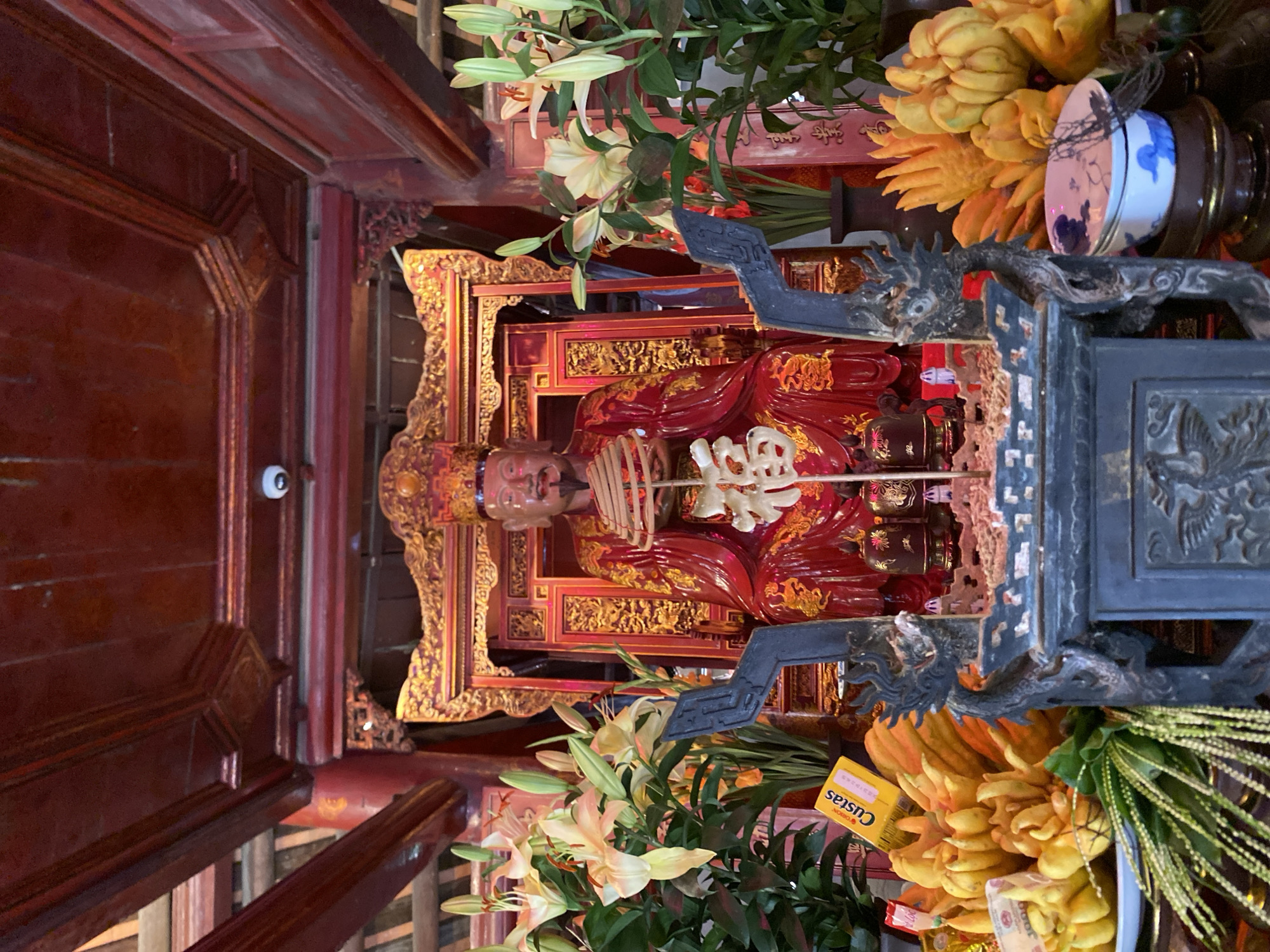 The image depicts the interior of the Temple of Literature in Hanoi, Vietnam. In the center, a statue of a historical figure, likely a scholar or emperor, is seated on an ornate, gilded throne. The figure is dressed in vibrant red robes adorned with gold embroidery, wearing a crown. They hold a symbolic object in their hands, with a decorative character inscribed on it.
Surrounding the throne are offerings, including a large cluster of Buddha's hand fruits on the left and right, as well as a bouquet of lilies. A small yellow box with the brand "Custas" is visible among the offerings on the left. In the foreground, a dark, intricately carved incense burner with dragon motifs is positioned. Behind the statue, a red and gold decorated backdrop is present. The ceiling is made of dark wood, with visible beams and decorative elements. The overall atmosphere is one of reverence and cultural significance.