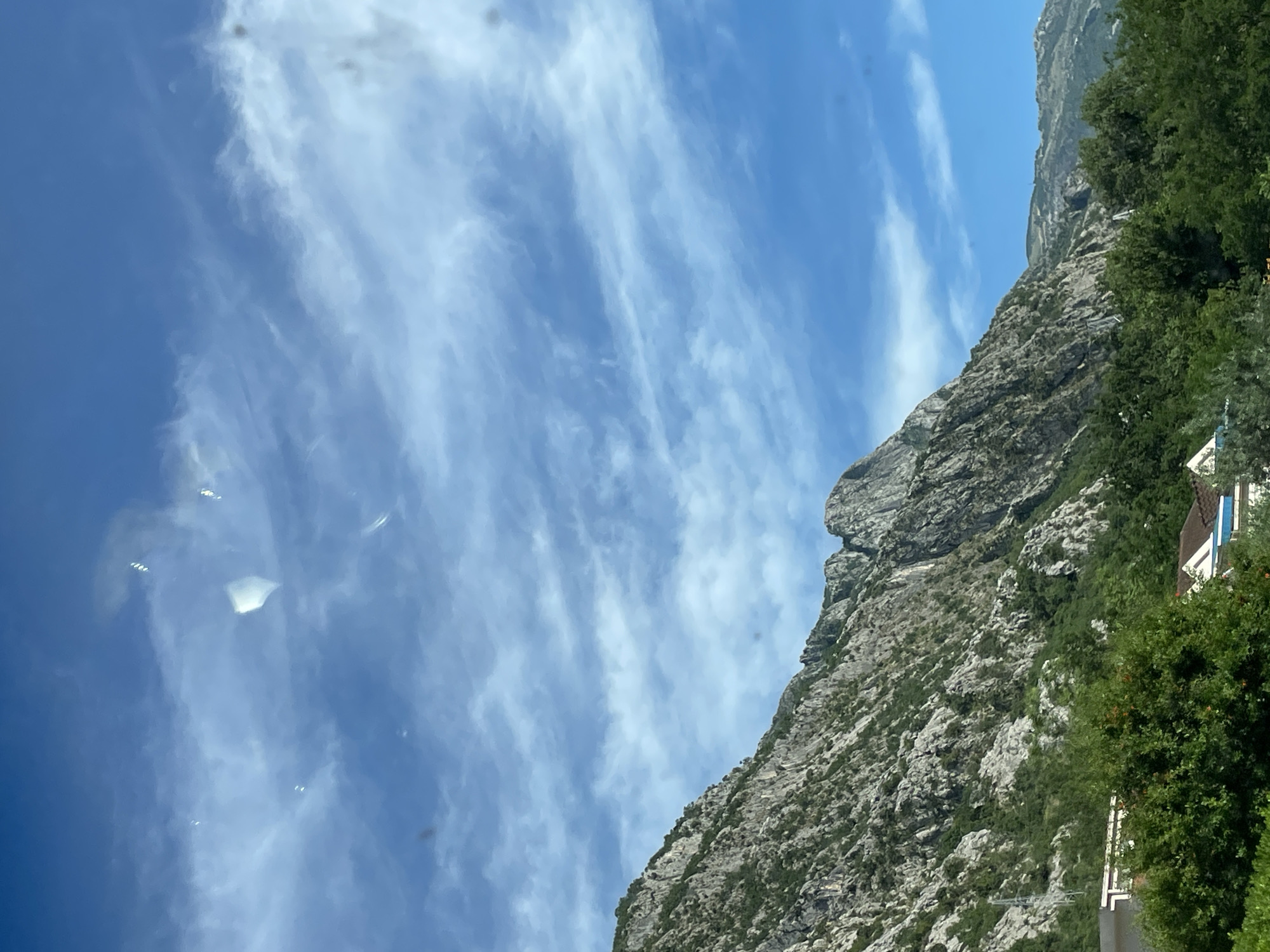 The image shows a rocky mountain in Dobrota, Montenegro, with a clear blue sky dotted with streaky, cirrus-like clouds. The mountainside is sparsely covered with greenery. A building with a brown roof and white walls is partially visible, obscured by trees.