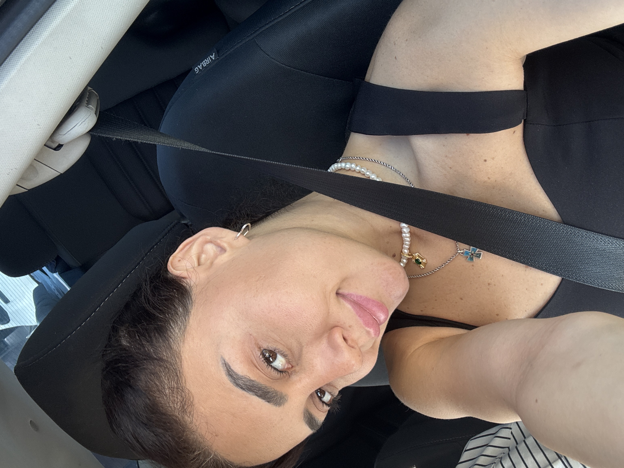 The image shows a woman in a car, likely in Tbilisi, Georgia, based on the prompt. She is in the driver's seat, with a seatbelt fastened across her. The woman has dark hair pulled back and is wearing a black top and two necklaces, one with pearls and the other a silver chain with a blue cross pendant. She is looking towards the camera with a slight smile. The car interior is visible, including the headrest of the seat and part of the dashboard. The lighting suggests it is daytime.