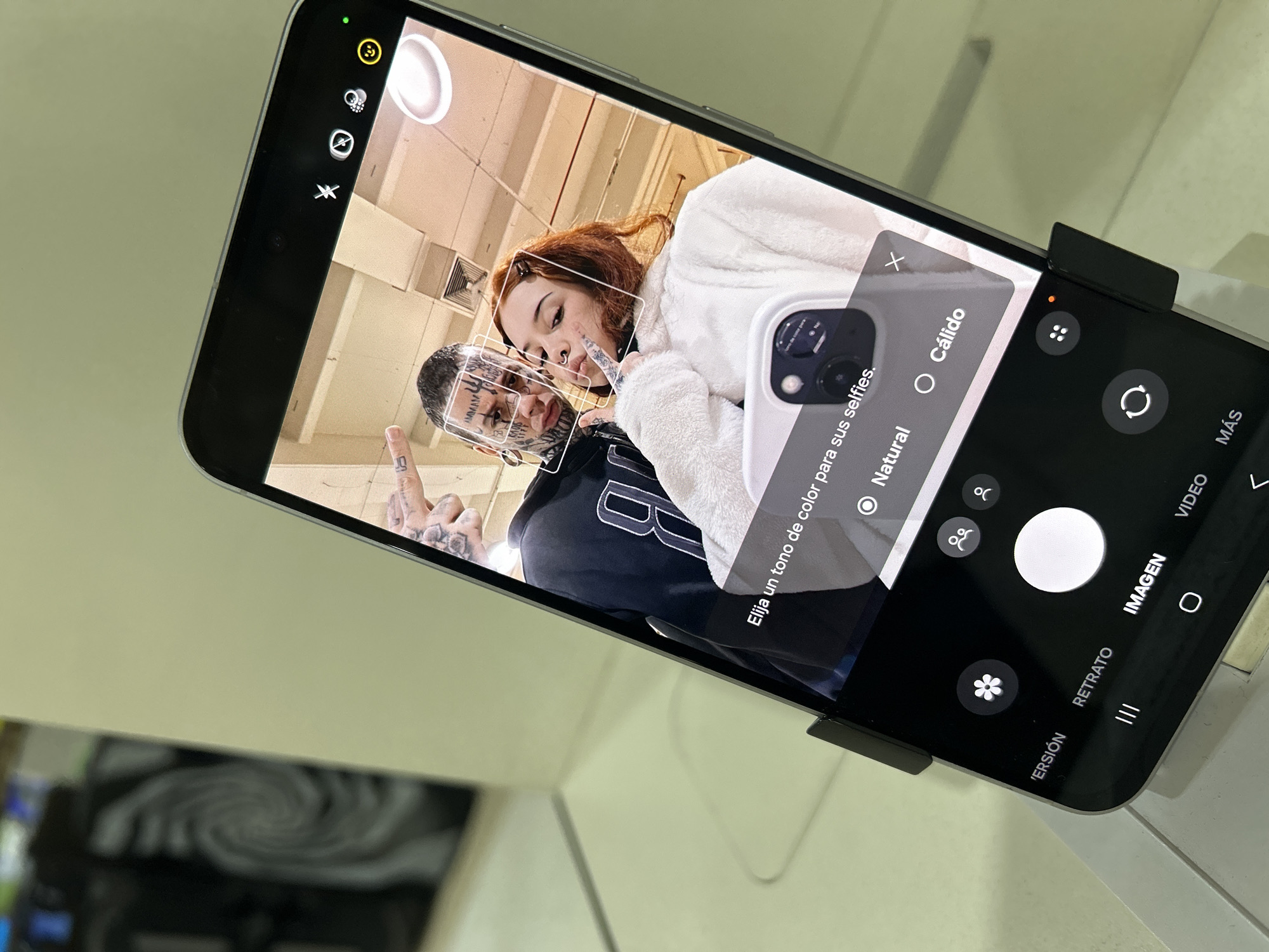 A smartphone displays a selfie of a man and a woman, both heavily tattooed and posing closely. The man is wearing a dark-colored shirt with white lettering, and the woman has red hair and is wearing a fluffy white garment. The phone screen shows the camera app with options for color tone selection, including "Natural" selected. The phone is held in a black stand. The background is light-colored and blurry.