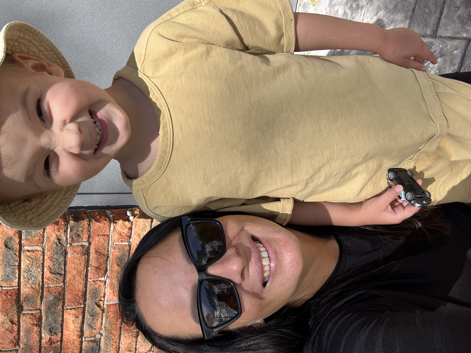 The image is a selfie taken by two people, a child and an adult, outdoors. The child is in the upper part of the frame, wearing a light-colored bucket hat and a yellow t-shirt. They are smiling widely, with their eyes closed. The adult is in the lower part of the frame, wearing sunglasses and a black t-shirt. They are also smiling.
In the background, there is a grey wall on the left and a brick wall on the right. The ground appears to be paved with stone tiles. The lighting suggests it is daytime, likely sunny, casting shadows.
The adult's hand is visible, holding a small black toy car. The overall mood of the image is happy and casual, suggesting a relaxed moment between the two subjects. There is no visible text in the image. The location context provided is Chaikovsky, Russia.