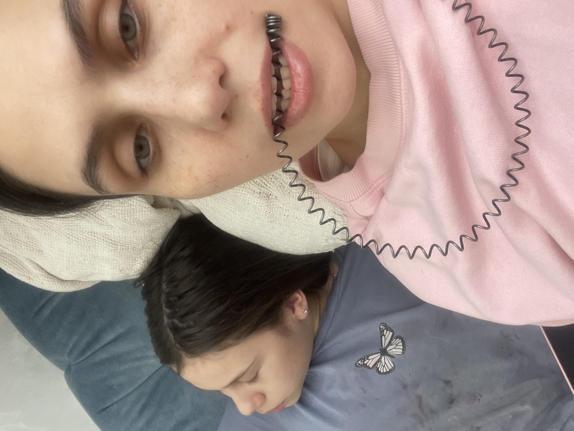 An adult female is captured in a close-up selfie indoors, looking directly at the camera. She has dark hair, green eyes, and light freckles. She wears a light pink long-sleeved top. A dark coiled metal spring is held horizontally between her lips, extending onto her chest. Behind her and to the left, a younger female with dark hair, styled with visible braids, lies on her side, eyes closed, facing away from the camera. She wears a light blue t-shirt featuring a white and black butterfly graphic on the left chest, and small silver stud earrings are visible in her earlobes. The light blue t-shirt has several dark smudges. Both individuals are positioned against soft furnishings, including a teal-blue upholstered surface and a cream-colored knitted textile. The specific location within the city of Izhevsk, Russia, cannot be determined from the image.