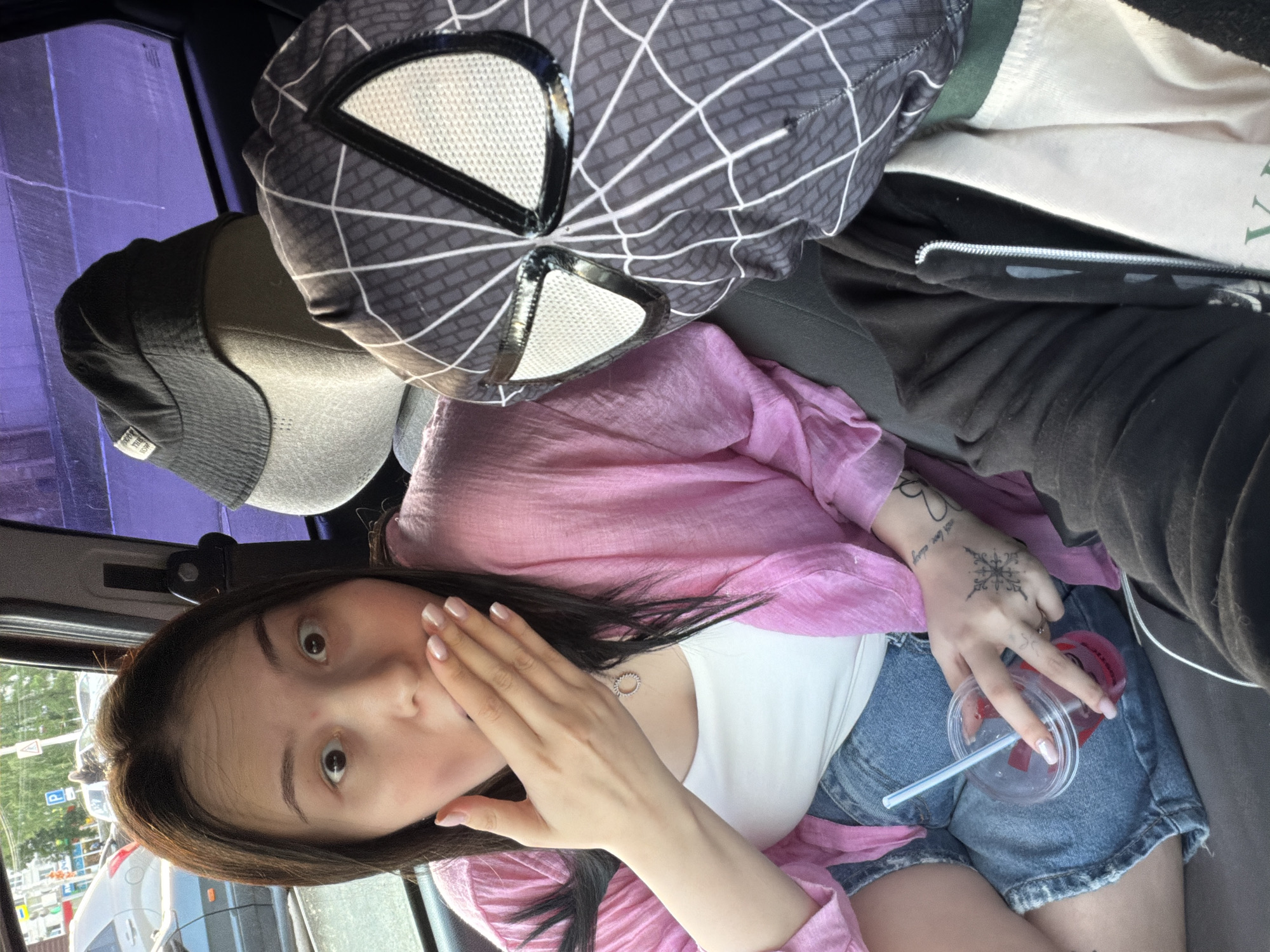 The image captures a young woman seated in what appears to be the passenger seat of a car, looking directly at the camera with a wide-eyed, surprised expression, her left hand partially covering her mouth. She has dark hair, fair skin, and is dressed in a white tank top, a sheer pink jacket, and denim shorts. Her fingernails are manicured with a light polish. Tattoos are visible on her left hand and wrist, including a distinctive snowflake-like pattern and partially legible text that appears to say "never love always." She holds a clear plastic cup with a red beverage and a blue straw in her right hand, which rests on her lap.
Behind the woman's head, a black Spider-Man themed headrest cover or pillow, featuring the characteristic white eyes of the black suit, is prominently displayed. Next to it, a dark grey cap is visible, along with various items of clothing in the background, including a black zippered hoodie and a light-colored garment with partially visible green text, possibly reading "WAY."
The car's side window, which has a noticeable purple tint, reveals a bright, clear daytime street scene outside. Trees, buildings, and other vehicles are visible, indicating an urban setting in Chaikovsky, Russia. The overall impression is a candid moment of surprise captured inside a vehicle during daylight hours. Visible text includes the partial text on her hand tattoo, and indistinct text on the Spider-Man cover and the cap's tag.