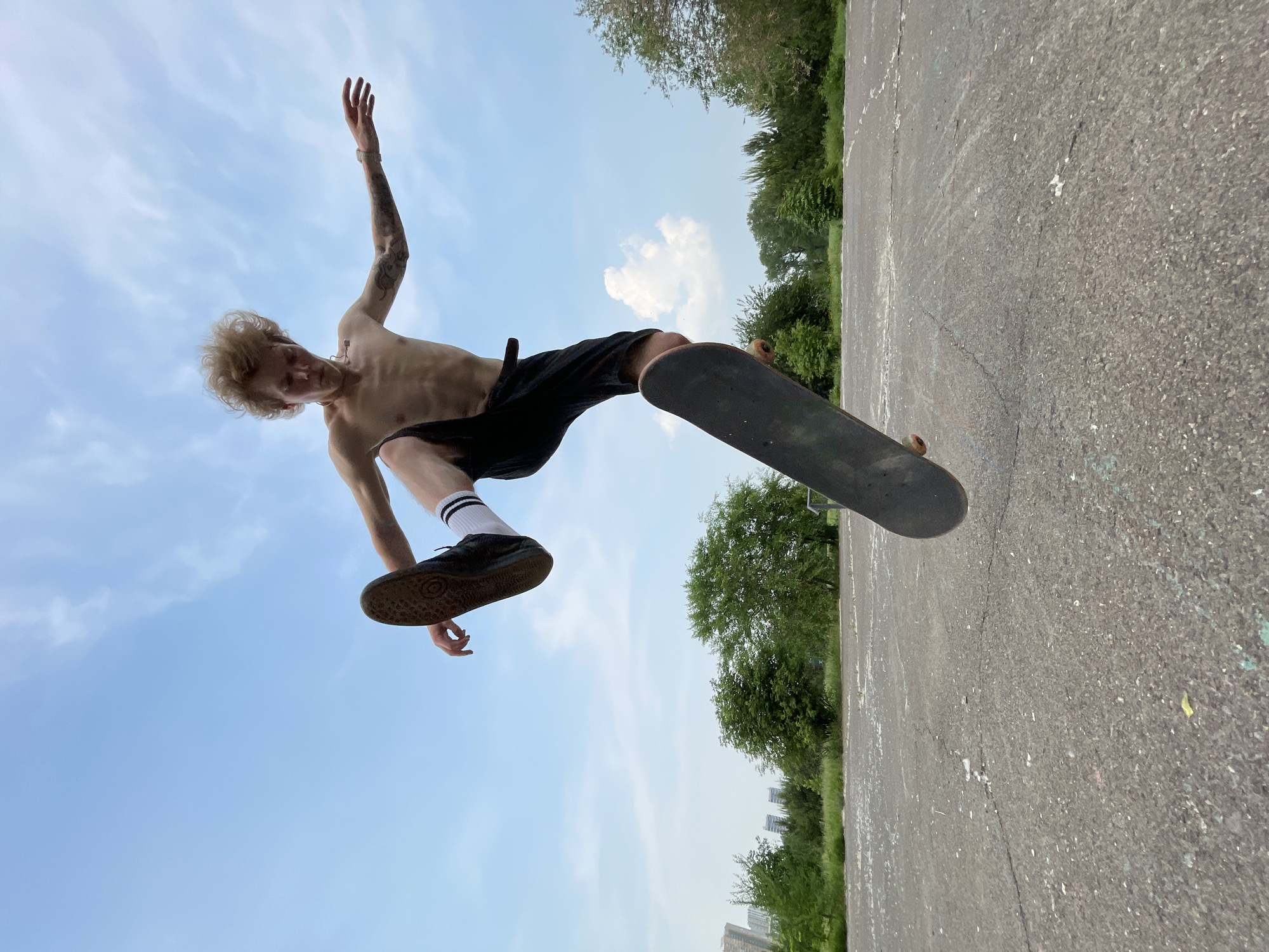 A young, shirtless male with blond hair and tattoos is captured mid-air performing a skateboarding trick. He is wearing black shorts with white striped socks and dark sneakers. The skateboard is airborne beneath him, oriented diagonally. The background consists of an outdoor asphalt area with scattered debris, leading to a grassy area with trees and distant buildings. The sky is bright blue with scattered white clouds. The image appears to have been taken in an urban park or designated skateboarding area, possibly in Krasnoyarsk, Russia, given the architectural style of the distant buildings and the general landscape.