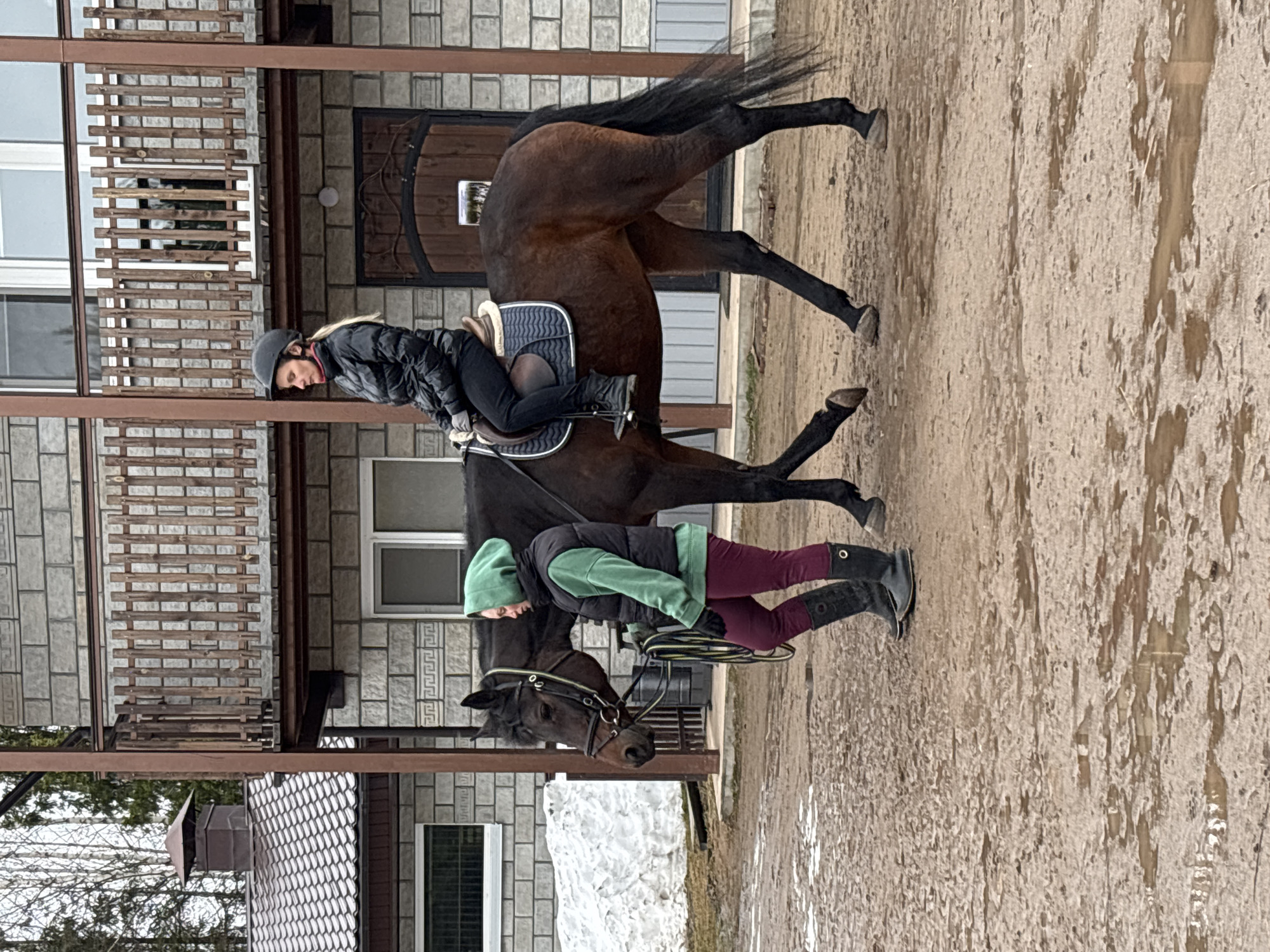 The image depicts two horses and riders in an outdoor setting. One rider is mounted on a brown horse, wearing a black jacket, black helmet, and black pants. The horse appears to be trotting, with its tail raised. Another individual is standing next to another horse, wearing a green hoodie, black vest, and burgundy pants, holding the horse's reins. A building with stone exterior and metal beams is in the background. The ground appears muddy or wet. The image was taken in Русский Вожой, Russia.