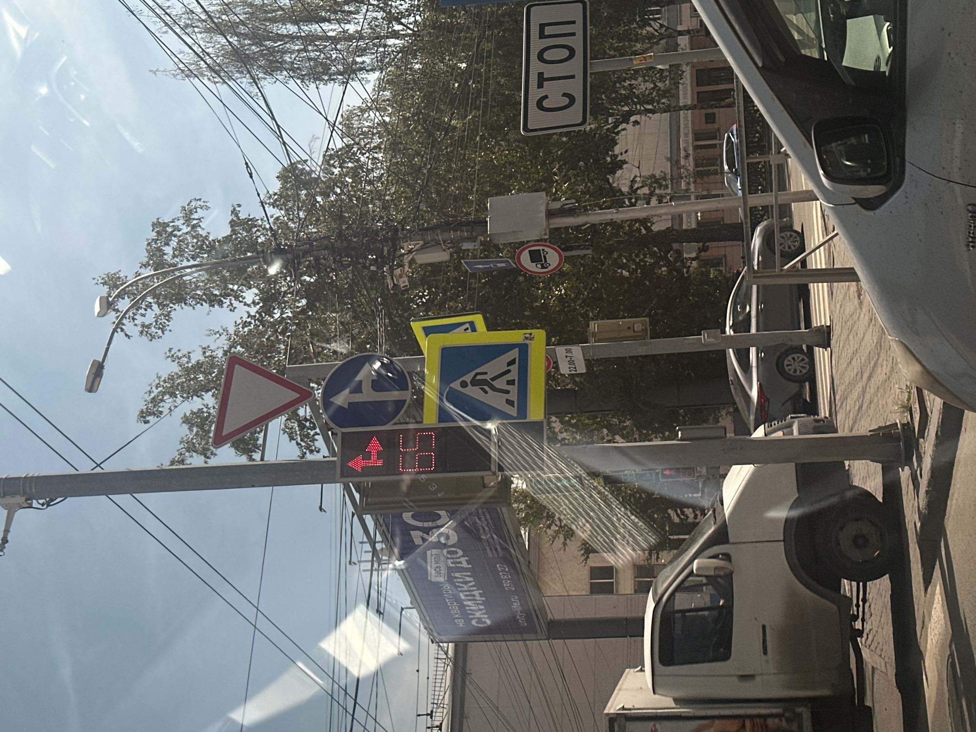 The image, taken from inside a vehicle, depicts a sunny daytime street scene, rotated 90 degrees clockwise. The foreground is dominated by reflections on a window or windshield.
Numerous road signs and traffic lights are visible. A large vertical "СТОП" (STOP) sign is prominently displayed at the top right. Below it, a circular red-bordered sign with a white truck symbol indicates a restriction, possibly for cargo vehicles, with a smaller rectangular sign underneath showing a time restriction "22.00-7.00".
Further down and to the left, a triangular yield sign (give way) is visible. Below it, a blue circular sign with a white arrow indicates a left turn. Adjacent to it is a blue square sign with a walking person, signifying a pedestrian crossing. A traffic light below these signs shows a red countdown "15" and a red left arrow, indicating that vehicles turning left need to wait.
In the right foreground, part of a white truck or van is visible, alongside other parked or moving cars in the middle ground, including a gray sedan and a blue car. Overhead wires and cables crisscross the sky. Lush green trees are abundant, suggesting a leafy street or park area. Buildings are discernible in the background.
A large billboard is visible below the traffic lights, displaying Cyrillic text including "на квартиры весь май" (for apartments all May), "СКИДКИ ДО" (DISCOUNTS UP TO), and a website "unity-life.ru" along with a phone number "230 97 27". The bright sunlight and clear sky indicate good weather conditions.