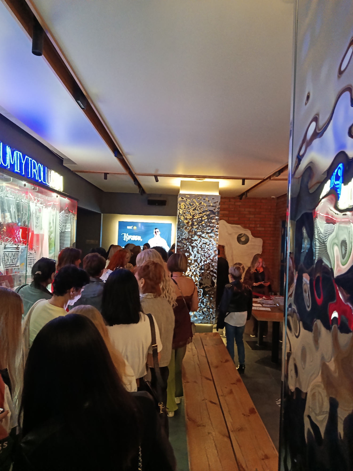 The image shows a queue of people inside a shop in Moscow, Russia. The name of the shop is "MUMIY TROLL", indicated by a blue neon sign. The shop is selling merchandise, including clothing and accessories, displayed behind glass. A screen displays an image of a man in a white shirt. A woman stands at a table with books, presumably selling or signing them. The interior design includes a brick wall, a decorative metallic structure, and a long wooden bench. The people are standing in line, facing forward, waiting their turn.