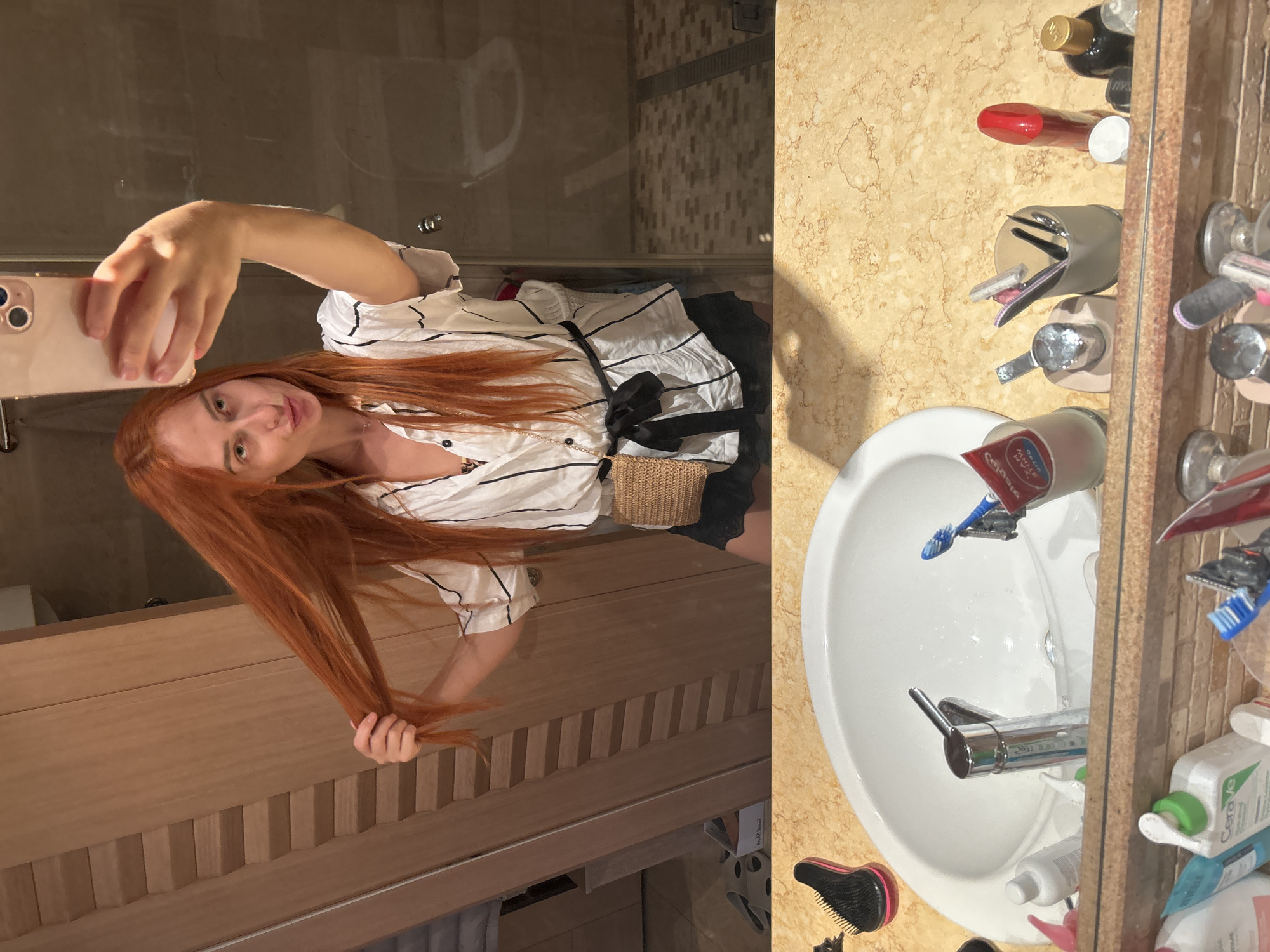 A young woman with long, fiery red hair takes a mirror selfie in a bathroom. She is wearing a white shirt with black pinstripes and a black ribbon tied at her waist. In her right hand, she holds a rose gold smartphone, angled to capture her reflection. Her hair cascades over her shoulders and down her arm, and she is pulling a section of it with her left hand.
The bathroom appears to be well-lit, suggesting it's daytime, though the exact time is not discernible. The scene is divided into two main parts by the reflection in the mirror. To the left, the woman is in what looks like a shower area with glass doors and tiled walls. To the right, the vanity area is visible, featuring a white sink with a chrome faucet. Various toiletries are scattered around the sink, including a blue toothbrush, a tube of toothpaste, and several bottles of skincare products. A small, woven straw bag with a black ribbon is also visible near the woman's waist in the reflection.
The vanity countertop is made of a marbled yellow and beige material. The background behind the woman shows wooden cabinetry with a ribbed texture. The overall impression is of a well-appointed bathroom, possibly in a hotel or a modern apartment, in Budva, Montenegro. The woman's expression is a gentle smile, conveying a relaxed and perhaps playful mood. Visible text includes "Colgate" on a tube of toothpaste.