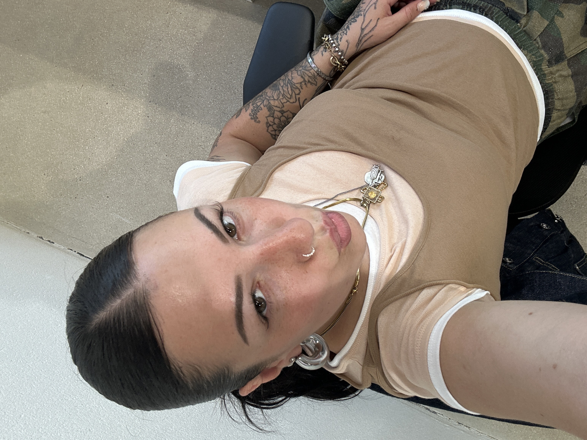 A woman is sitting on a black chair in a brightly lit room with a tan floor. She is wearing a tan tank top over a peach t-shirt with white trim, along with camouflage pants. She has a tattoo on her left arm, and several necklaces around her neck. Her hair is pulled back. There are silver hoop earrings in her ears and a silver nose ring in her nostril. Part of a denim jacket is visible near the chair. The location in the city of Paris, France is not discernable from this image.
