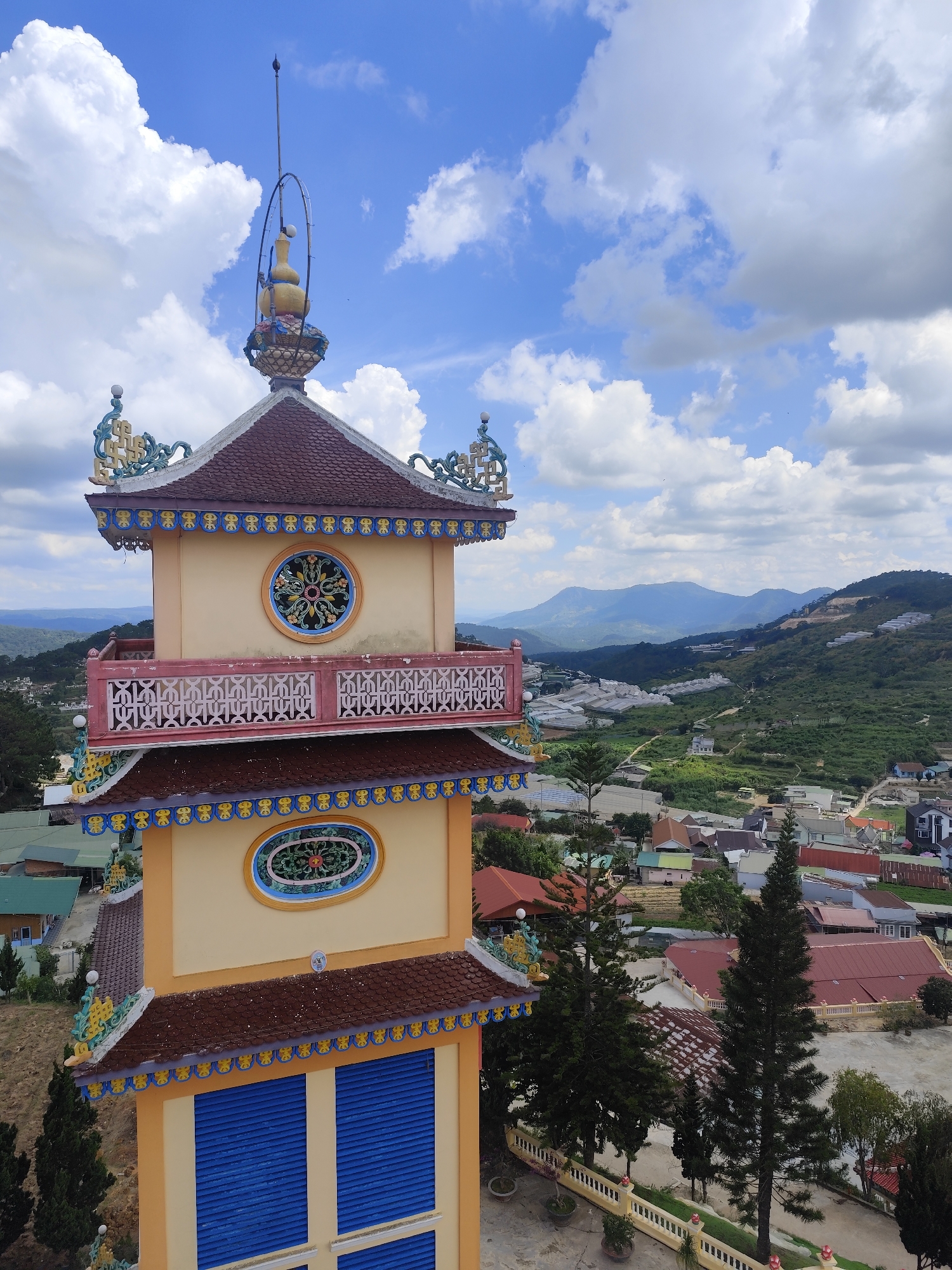 This is a high-angle, eye-level, medium shot of a multi-tiered pagoda in Lộc Quý, Vietnam. The pagoda is a tall, slender structure painted in shades of yellow, orange, and blue, with ornate decorative elements and tiled roofs. The top of the pagoda features a decorative finial with a gourd-like shape. Below the finial are intricate blue and yellow trim details and a circular window with an ornate floral design. The pagoda has a balcony with a pink railing featuring an intricate cutout pattern. Further down, another circular window with a green floral design is visible. The lower section of the pagoda features large blue slatted window coverings.
The pagoda is situated on a hillside overlooking a town with a mix of residential buildings and greenhouses. The town extends into the background where rolling, green hills and mountains are visible under a bright blue sky filled with fluffy white clouds. Several tall, dark green pine trees are positioned in the foreground and middle ground, to the right and left of the pagoda, casting shadows on a paved courtyard area. The scene appears to be a bright, sunny day. There are no people visible in the image.