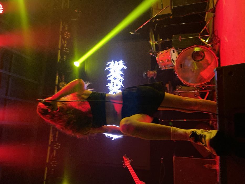 A performer in a black top and shorts bends backward over a microphone stand. Long, wavy, reddish-brown hair nearly touches the stage. Behind the performer, a drummer is visible in a black shirt, and drums are lit with red light. Stage lights are directed toward the performer, with red and yellow light dominating the scene. A bass guitar rests on the stage. A decorative light fixture stands between the performer and the drummer. The image is slightly rotated.
