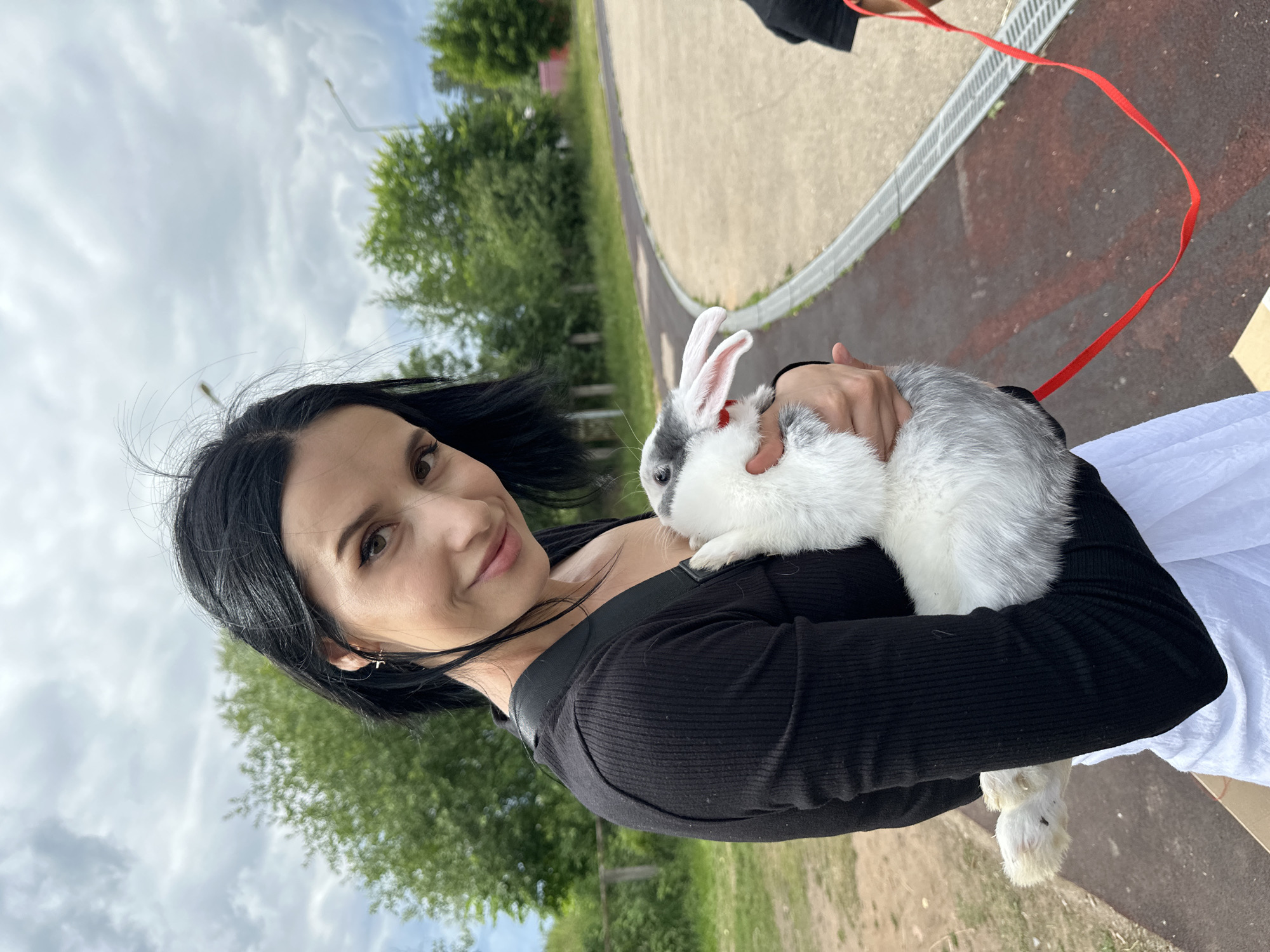 A young woman with dark hair and makeup is holding a small white and gray rabbit with pink ears in her arms. She is wearing a black long-sleeved shirt and a white skirt. The woman is smiling at the camera. The rabbit is held close to her chest. A red leash is attached to the rabbit's neck and extends out of frame. The background shows trees and a cloudy sky. The location appears to be an outdoor area, possibly a park or a farm, given the presence of a fence in the background and a running track with a drainage grate. The overall setting does not specifically indicate the city of Chaikovsky, Russia.