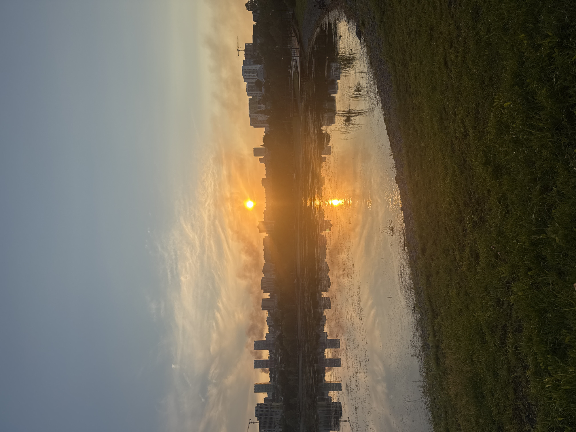 This is an outdoor, eye-level shot taken at dusk of a lake with a city skyline in the background. The sun is low in the sky, casting a warm orange glow across the scene and reflecting on the water. The water is calm and shows reflections of the buildings and the sky. The shoreline in the foreground is covered in grass and rocks. There are several tall buildings in the city, some of which appear to be under construction, indicated by a crane. There is also a plume of smoke rising from behind the buildings. The overall mood is peaceful, with the natural beauty of the sunset contrasting with the urban landscape. The image is likely taken at a lake in Ufa, Russia.