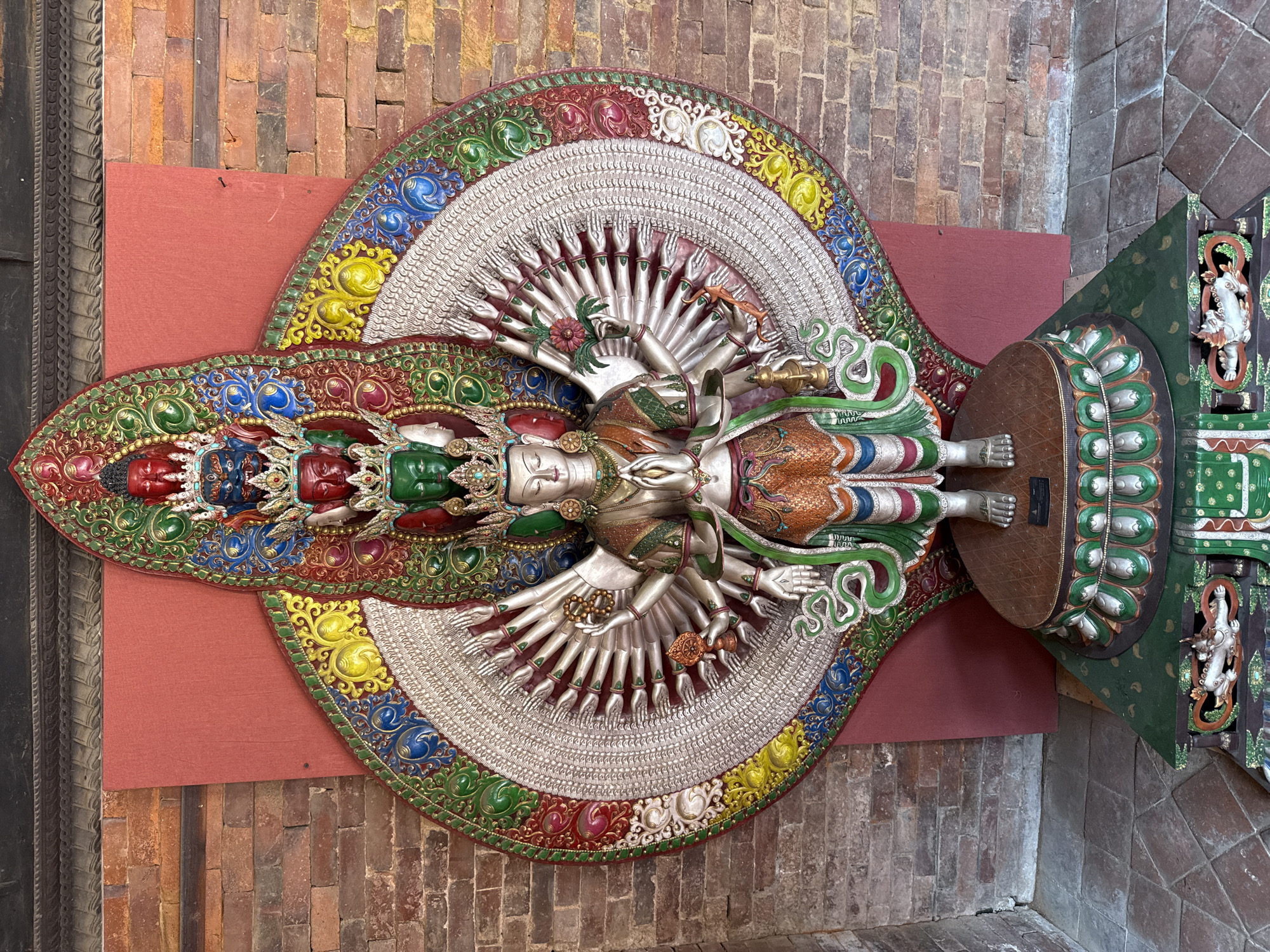 The image depicts a large, intricate sculpture of a deity, likely a Buddhist or Hindu figure, mounted on a wall in a building in the city of Lalitpur, Nepal. The sculpture is multi-armed, with a detailed and colorful head, torso, and legs. The hands radiate outwards from the body in a fan-like arrangement. The background of the sculpture is a semi-circular design with a textured, silver-colored interior and a decorative border with vibrant colors such as red, green, yellow, and blue. The wall behind the sculpture is constructed of red bricks, and there is a red panel behind part of the circular design. To the right of the figure is part of a dark green base platform embellished with similar colorful patterns. The overall setting suggests an interior display within a temple, museum, or another cultural location in Lalitpur.