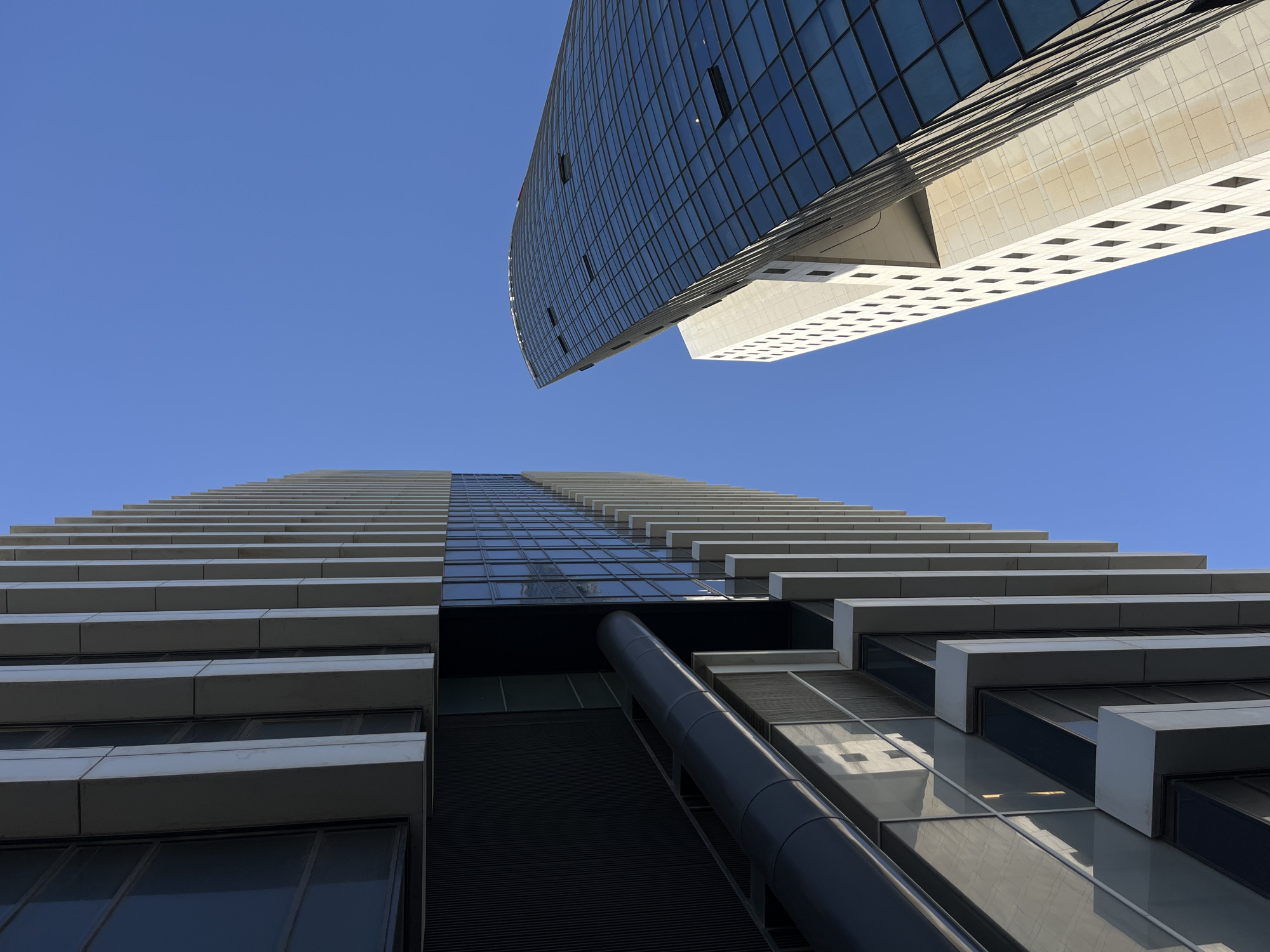 The image shows two modern buildings in Tel-Aviv, Israel, viewed from a low angle looking up at a clear blue sky. The bottom building is tall and narrow, with horizontal white rectangular protrusions extending from the glass and dark-colored facade. The top building has a curved glass surface with rectangular white protrusions on the bottom section. There are no people or signs of human activity visible in the scene.
