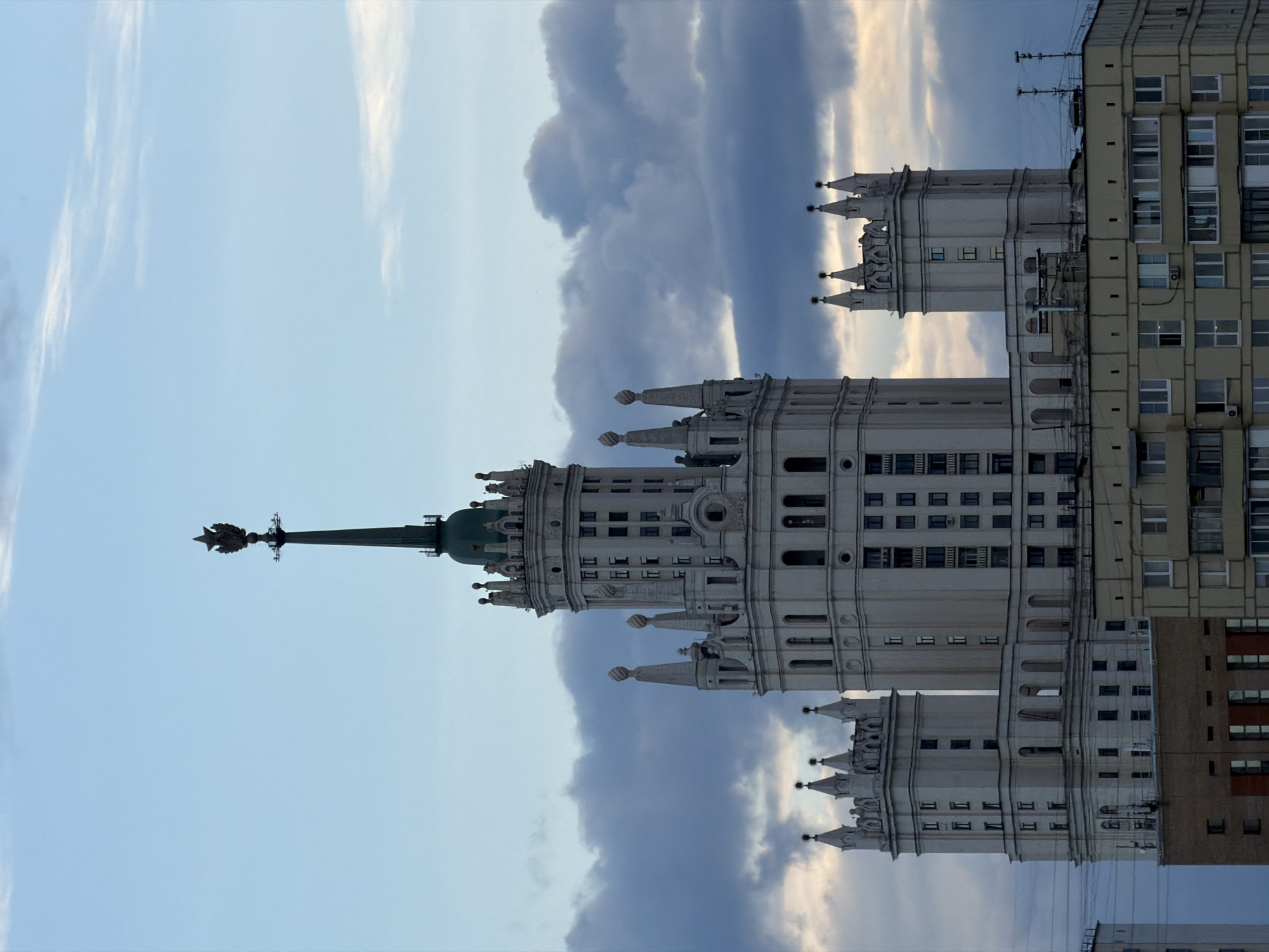 The image shows a tall, ornate building with a spire topped by a star against a cloudy sky. The building is white with multiple towers and intricate detailing. Adjacent to it is a more modern, rectangular building with rows of windows. The location is likely a city in the Soviet Union or former Soviet Union due to the architectural style of the ornate building. The photograph is taken at an angle, giving the impression that the buildings are leaning.