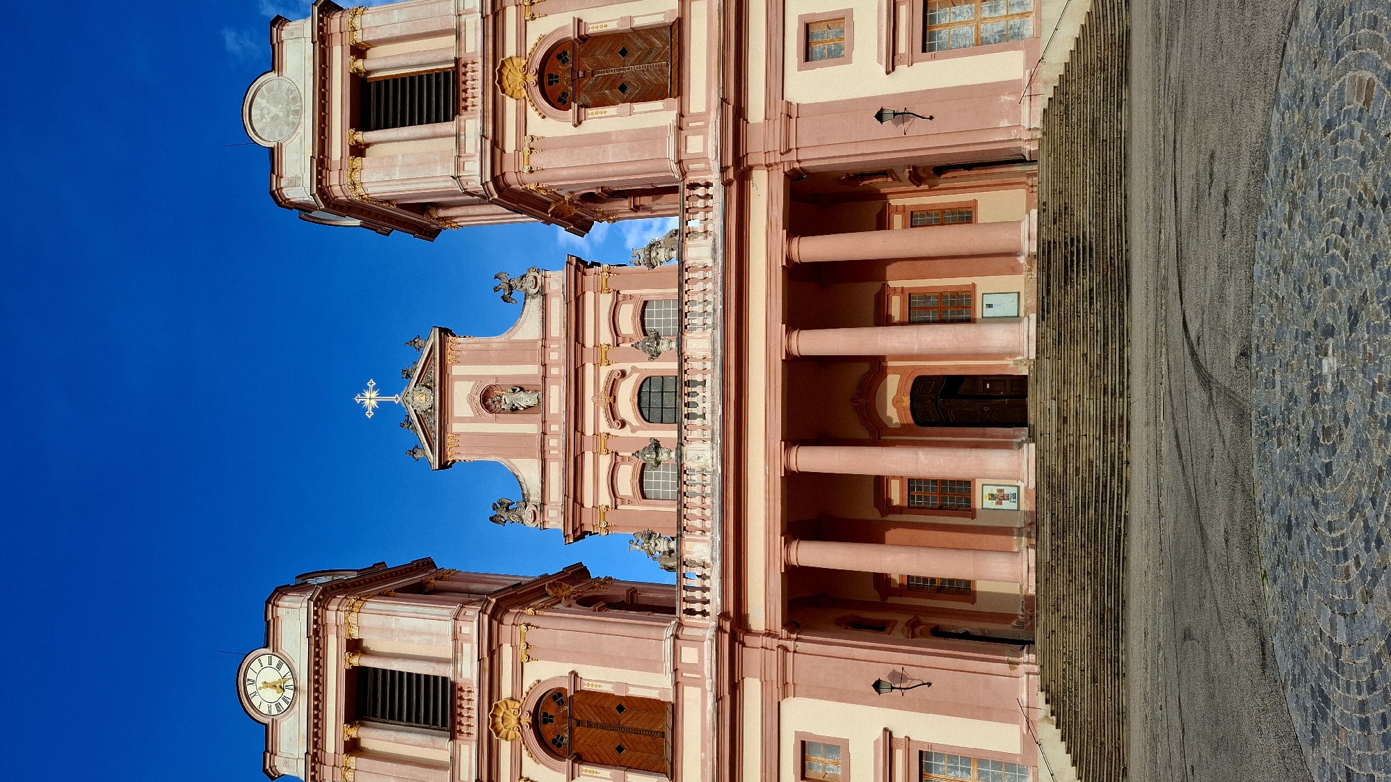 The image depicts the facade of Göttweig Abbey, a Benedictine monastery located in Furth bei Göttweig, Austria. The abbey is characterized by its Baroque architecture, with a central pink and cream-colored structure flanked by two towers. Each tower features a clock face and ornate detailing. A large set of stone stairs leads up to the main entrance, which is framed by colossal columns. The sky above is a clear, vibrant blue. There are no people visible in the image. The foreground shows a cobbled circular area and asphalt, suggesting a public square or entrance.