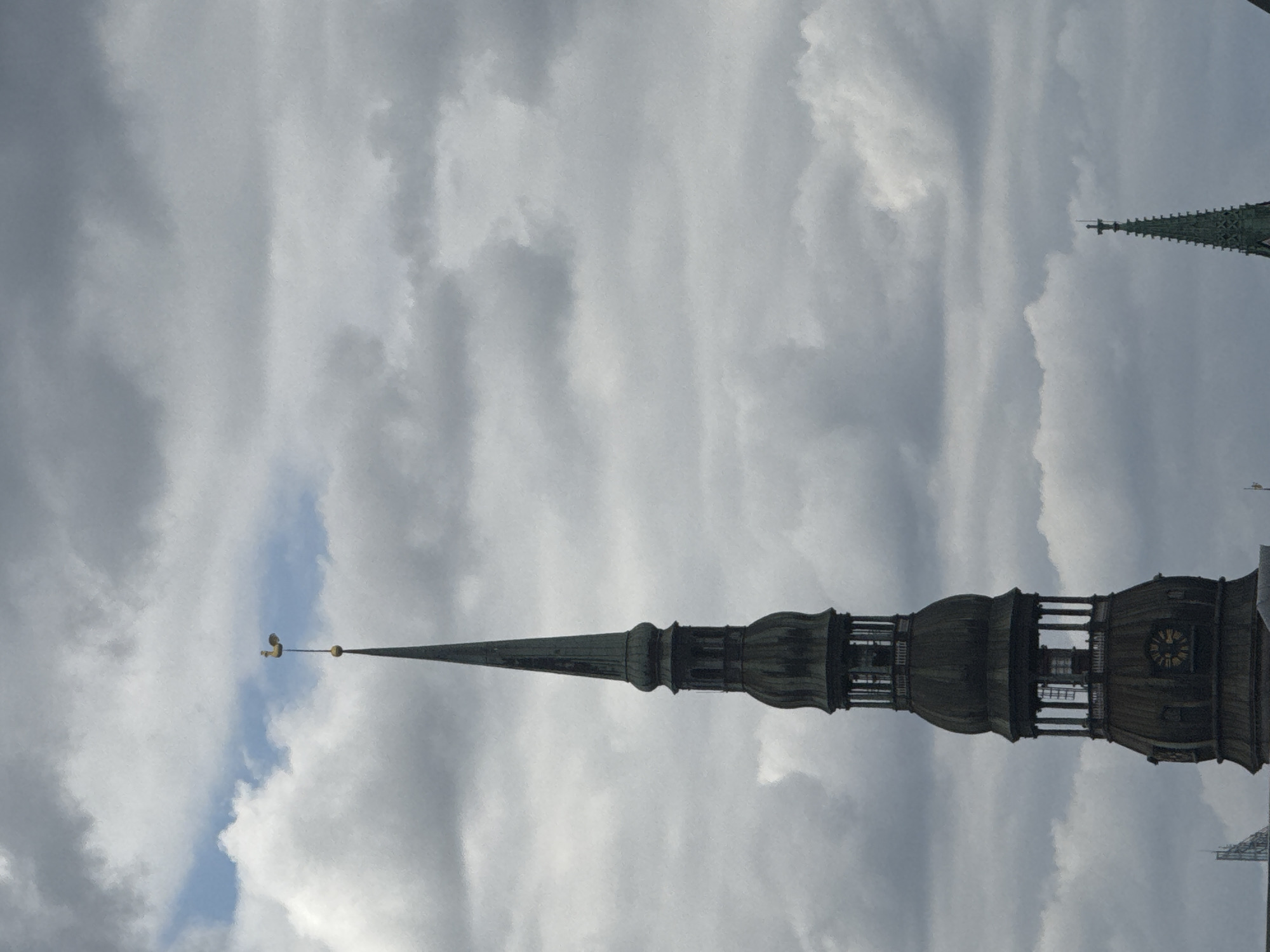 An architectural photograph presents a striking view of a tall, ornate church spire reaching into a cloudy sky. The main subject is a dark, multi-tiered Baroque-style tower, strongly resembling St. Peter's Church in Riga, Latvia, given the provided context. The spire tapers sharply to a slender point, crowned with a golden weather vane in the shape of a rooster.
The tower's design includes several levels featuring visible open sections or windows. A prominent clock face with golden numerals is discernible on one side of the belfry section, though the precise time is not clear. In the upper right background, partially obscured by clouds, the tip of another spire is visible, possibly Riga Cathedral.
The sky is largely covered by expansive grey and white clouds, indicating an overcast day. However, a distinct patch of blue sky can be seen to the left of the main spire, offering a contrast to the cloud cover. The overall lighting is diffused, suggesting daytime. No people or specific activities are depicted; the image focuses on the grandeur of the architecture against the dramatic sky.
