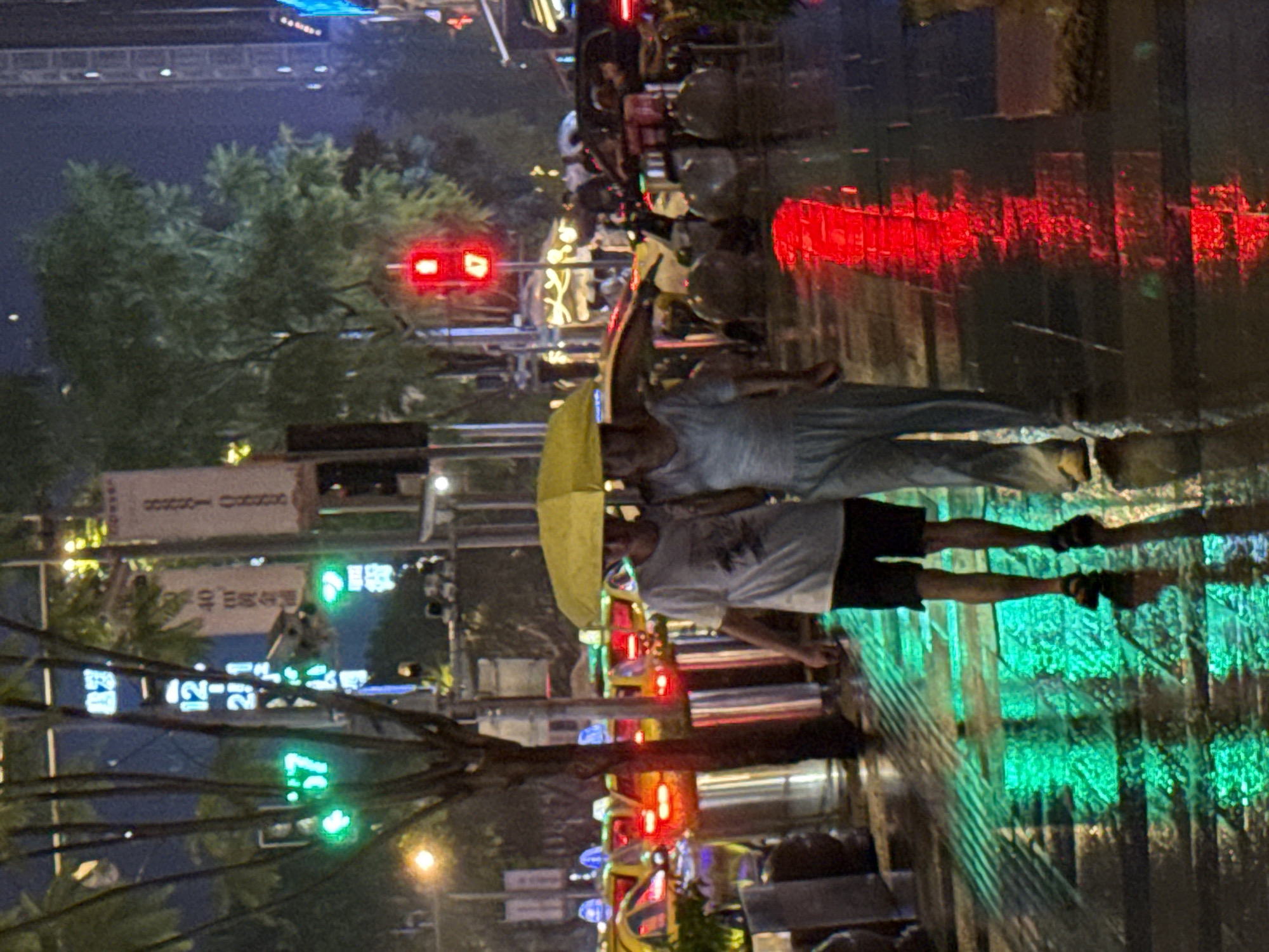 The image depicts a street scene at night in Liangjiang, China, during a rain shower. Two individuals are walking together under a yellow umbrella. The person on the left is wearing a white t-shirt and dark shorts, while the person on the right is wearing a light-colored jumpsuit. They are walking on a wet pavement that reflects the green and red lights from traffic signals and nearby illuminated signs. Yellow taxis are visible in the background, and trees line the street. The overall atmosphere is illuminated by streetlights and the reflections on the wet surfaces.