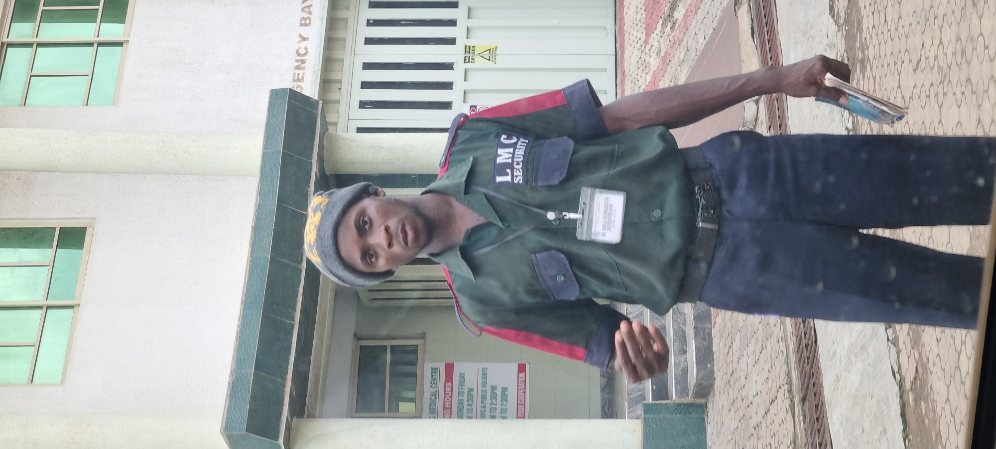 A man wearing a dark green security uniform with red and black accents on the sleeves and collar stands outside a building. He has a gray beanie on his head and a lanyard with an ID badge around his neck. The badge is visible and appears to have text and a photo on it. The man is holding a folded pamphlet or booklet in his right hand. The building behind him is white with green-tinted windows and a sign that reads "EMERGENCY BAY." Another sign on the building lists "LMC MEDICAL CENTRE OPERATING HOURS." The ground in front of the building is paved with interlocking stones and has some metal grates. The scene appears to be in a public area.