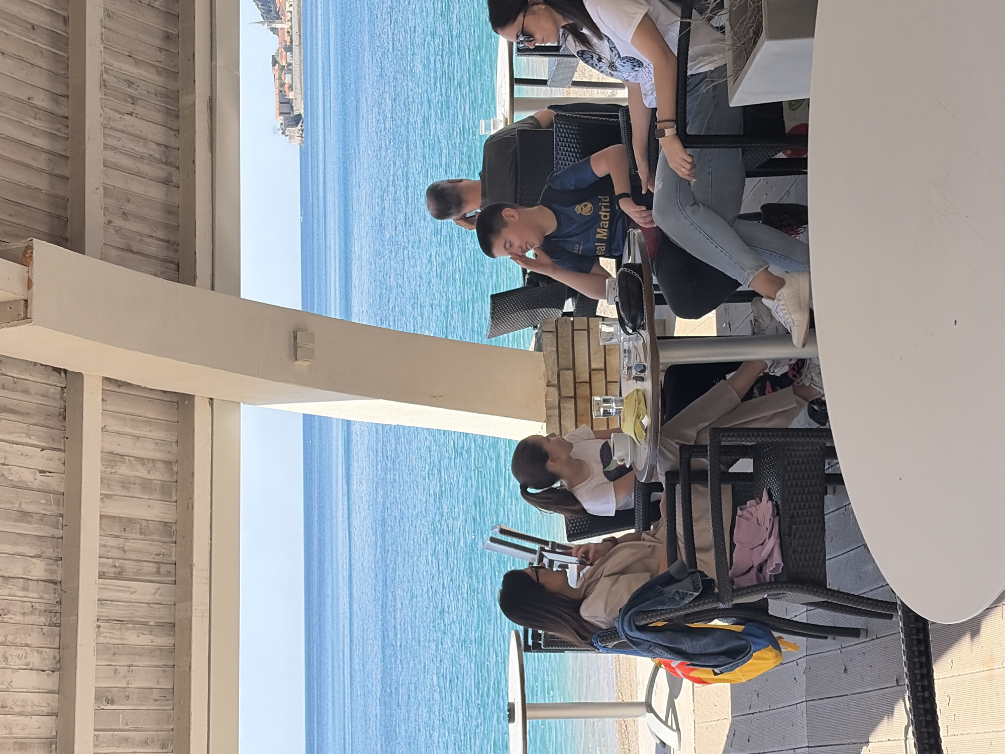 A group of people is seated at tables on the terrace of a restaurant overlooking the Adriatic Sea in Budva, Montenegro. To the left, a partly-visible roof covers the terrace. The turquoise sea stretches to the horizon. The city of Budva is visible in the distance on the upper left. Several people are seated at the tables, engaged in conversation. One is wearing a Real Madrid soccer jersey. Clothing and bags are draped over the chair backs. Glasses and other items are on the tables. The terrace is lined with tables and chairs.