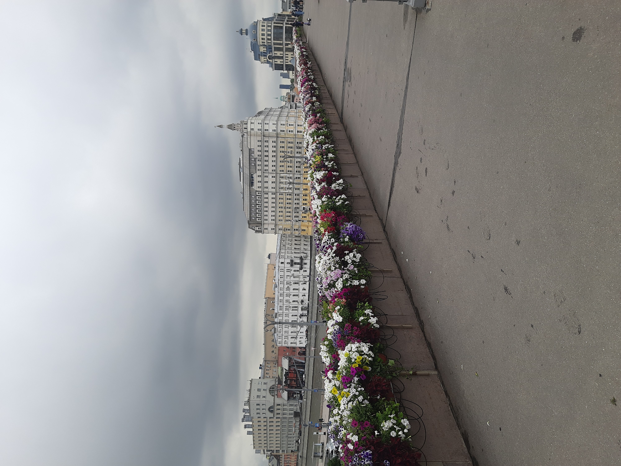 The image shows a wide, paved pedestrian walkway on a cloudy day. A long, rectangular planter box filled with colorful flowers runs along the left side of the walkway, creating a vibrant border. In the background, across a river (likely the Moskva River), several large, distinct buildings are visible. The architecture of these buildings appears to be a mix of historical and modern styles. One prominent building on the right has a distinctive dome. People can be seen walking on the walkway in the distance, and a scooter is parked near them. The overall scene suggests a public space within Moscow, Russia, likely a bridge or embankment, adorned with decorative floral arrangements.
