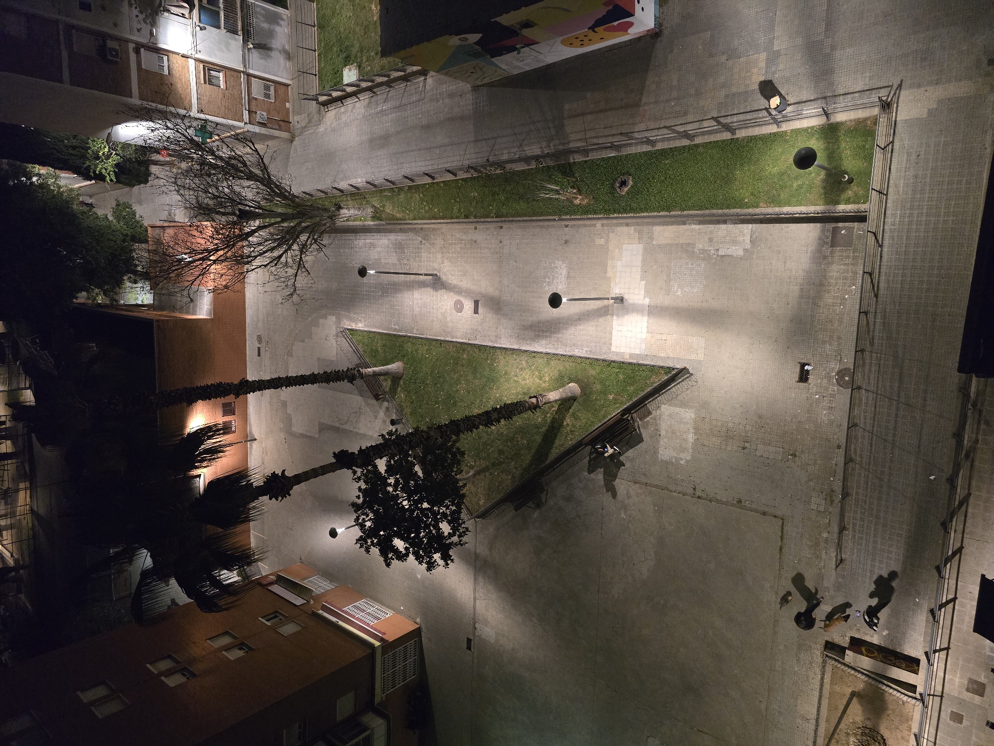 The image shows a top-down view of an urban area in Cornellà de Llobregat, Spain, at night. Several apartment buildings with visible windows are present. The streetscape includes paved areas, interspersed with triangular and rectangular grassy sections. Streetlights illuminate the scene, casting shadows. Trees, including palm trees and a bare tree, are scattered throughout the space. Several people and a dog are visible in the bottom right corner of the image. The scene is predominantly lit by artificial light, creating a contrast between light and shadow. There is a mural on one of the buildings on the top of the image.