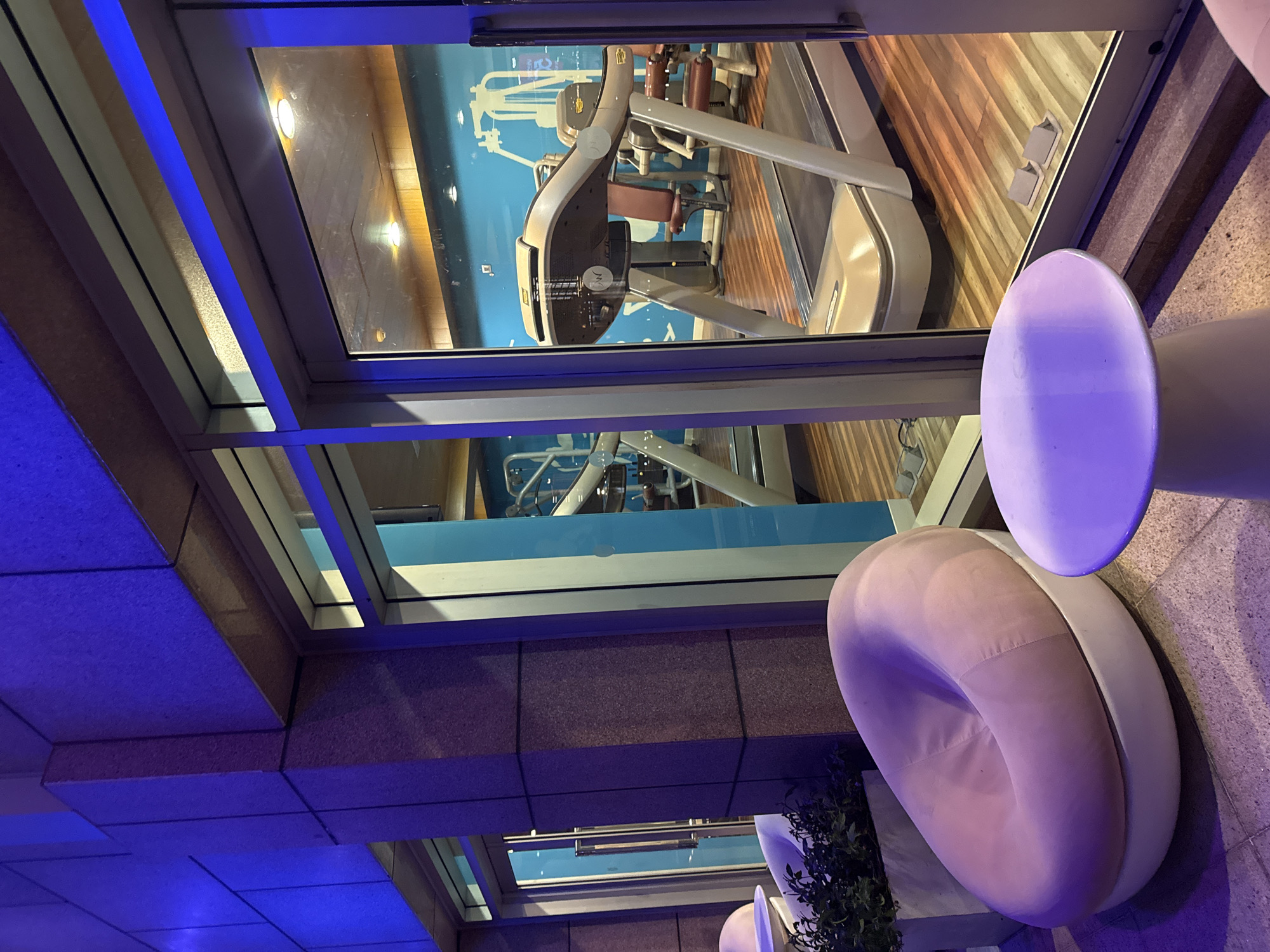 The image, oriented correctly, presents a modern indoor scene looking through a large glass window into a gym or fitness center. Inside the gym, visible through the window, are several pieces of exercise equipment, including treadmills and various weight machines with metallic frames and dark brown or red padded seats. The gym floor is made of light-colored wooden planks, and the interior is brightly lit by warm yellow overhead lights, contrasting with a light blue wall or mural in the background.
Outside the gym, to the right of the glass partition, a contemporary lounge area is illuminated by cool blue artificial lighting emanating from the ceiling and along architectural features. This area features a circular white table and a distinctive circular chair with a plush, light pink cushion set within a white frame. Both the table and chair reflect the blue ambient light, taking on purple and pinkish hues. Small potted plants are placed near the seating. The flooring in the lounge area appears to be a textured, light-colored tile.
No people are present in the image, and no specific activity is occurring, offering a static view of the architectural and interior design. The artificial blue and yellow lighting suggests it is nighttime or an interior space with controlled illumination. No legible text is visible on any objects. The overall impression is of a clean, sophisticated, and well-maintained facility, likely located in Đà Nẵng, Vietnam, as per the provided context.