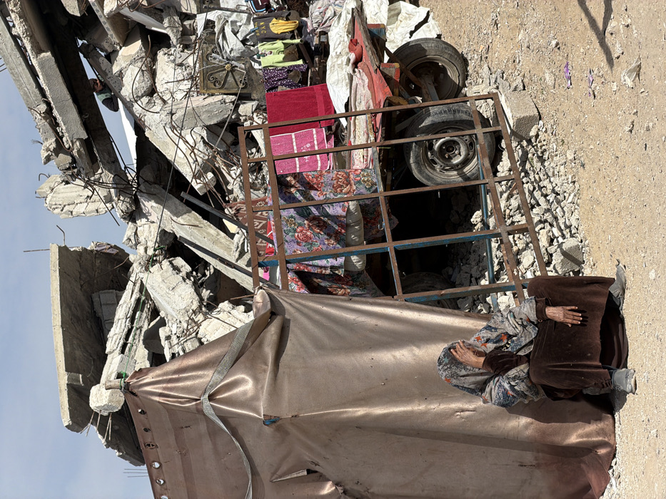 The image depicts a scene of extensive urban destruction in Jabalia, Palestinian Territory, under a clear daytime sky. In the foreground, an elderly individual is seated directly on the barren dirt ground, leaning against a makeshift shelter constructed from a shiny, coppery-brown tarp material secured with straps. This individual wears a multi-patterned head covering and a long, dark brown outer garment over a patterned shirt; their right hand is raised to their forehead, partially obscuring their face, while their left hand rests on their lap.
To the immediate right of the seated person, a rusty, rectangular metal frame, possibly from a bed or window grill, stands upright. Behind this frame, a red-framed utility cart with two visible wheels is positioned on uneven ground littered with small concrete debris. The cart is laden with various items, including patterned blankets and a red towel draped over its structure, and several articles of clothing (e.g., t-shirts, bags) hanging on a line or structure behind it. White tarpaulins are visible behind the cart.
The background is dominated by a massive pile of shattered concrete slabs and twisted rebar, indicating the remains of a demolished building. Amidst this rubble, a second individual, wearing a greenish top, is partially visible near the upper portion of the debris pile, appearing to be observing or navigating the destruction.