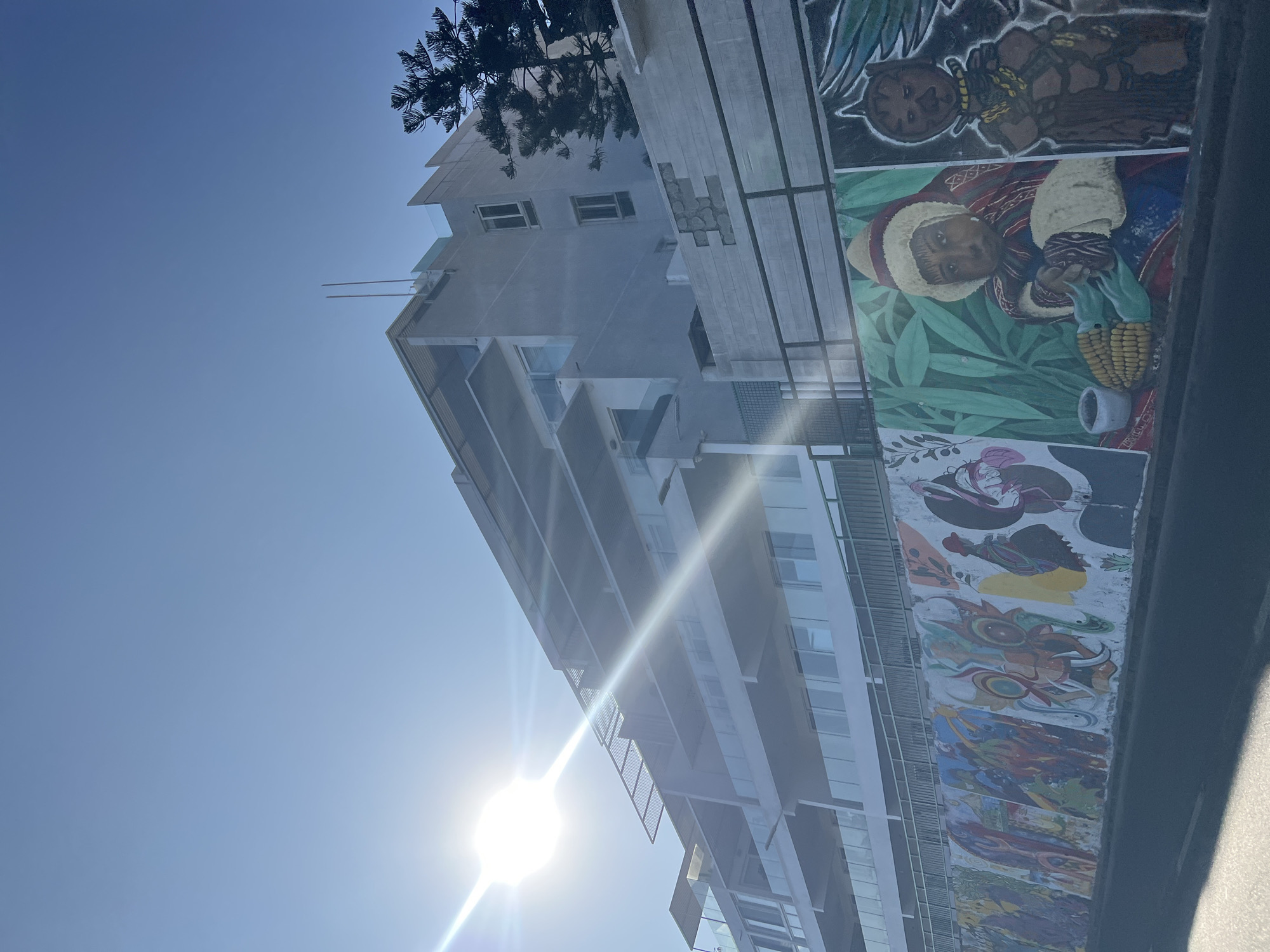 The image shows a tall building with a mural on its side in Arica, Chile. The mural depicts several images, including what appear to be indigenous people and other abstract designs. The building itself is modern and relatively high-rise. The sky is clear and blue, with the sun shining brightly, causing lens flare.