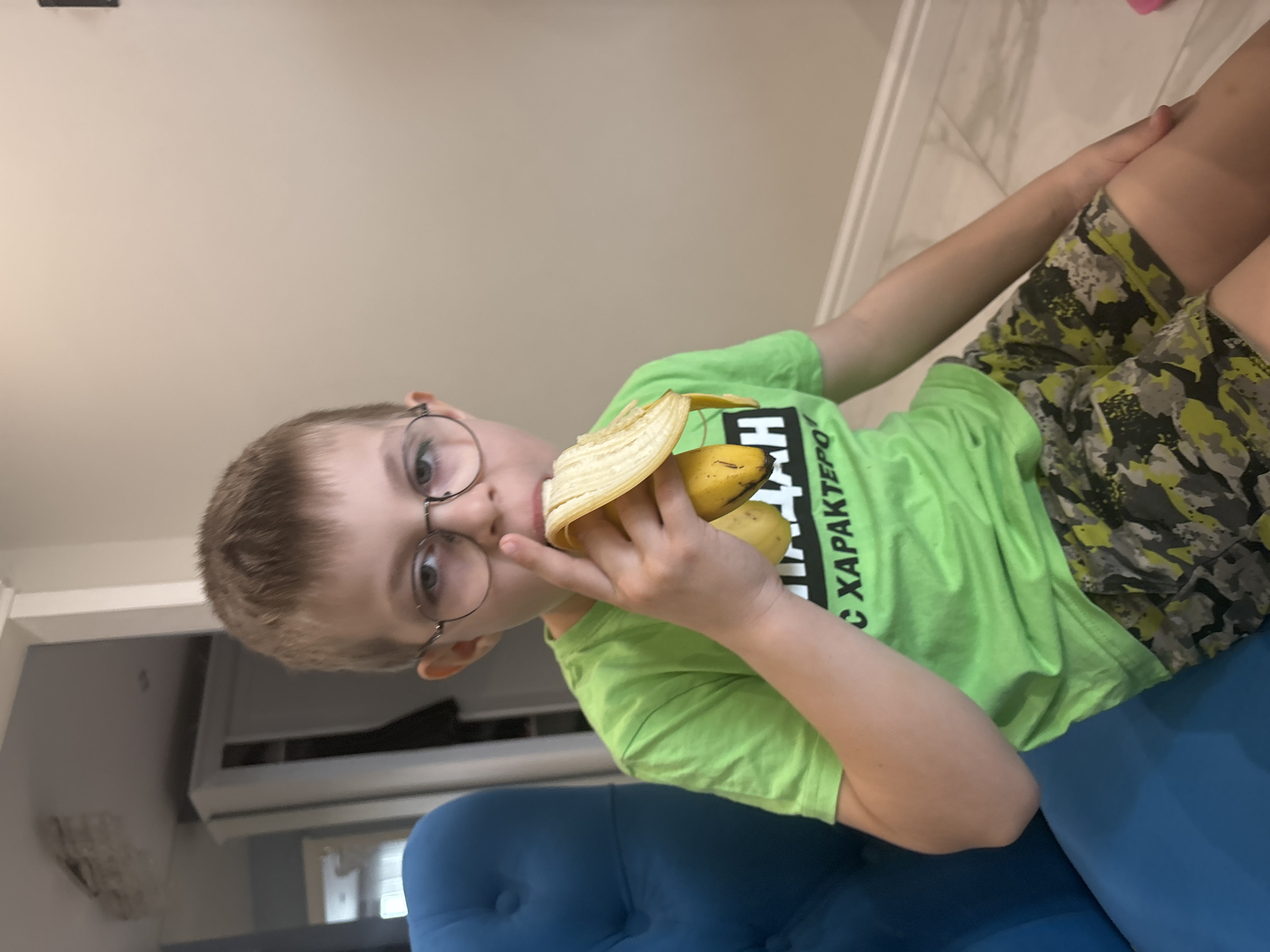 A young boy with short, light brown hair and round glasses is sitting on a blue, button-tufted chair. He is wearing a bright green t-shirt and camouflage patterned shorts. The boy is holding and eating a partially peeled banana. The background includes a white wall and a partially visible doorway with white cabinets. The location appears to be indoors. The city and place are unknown.