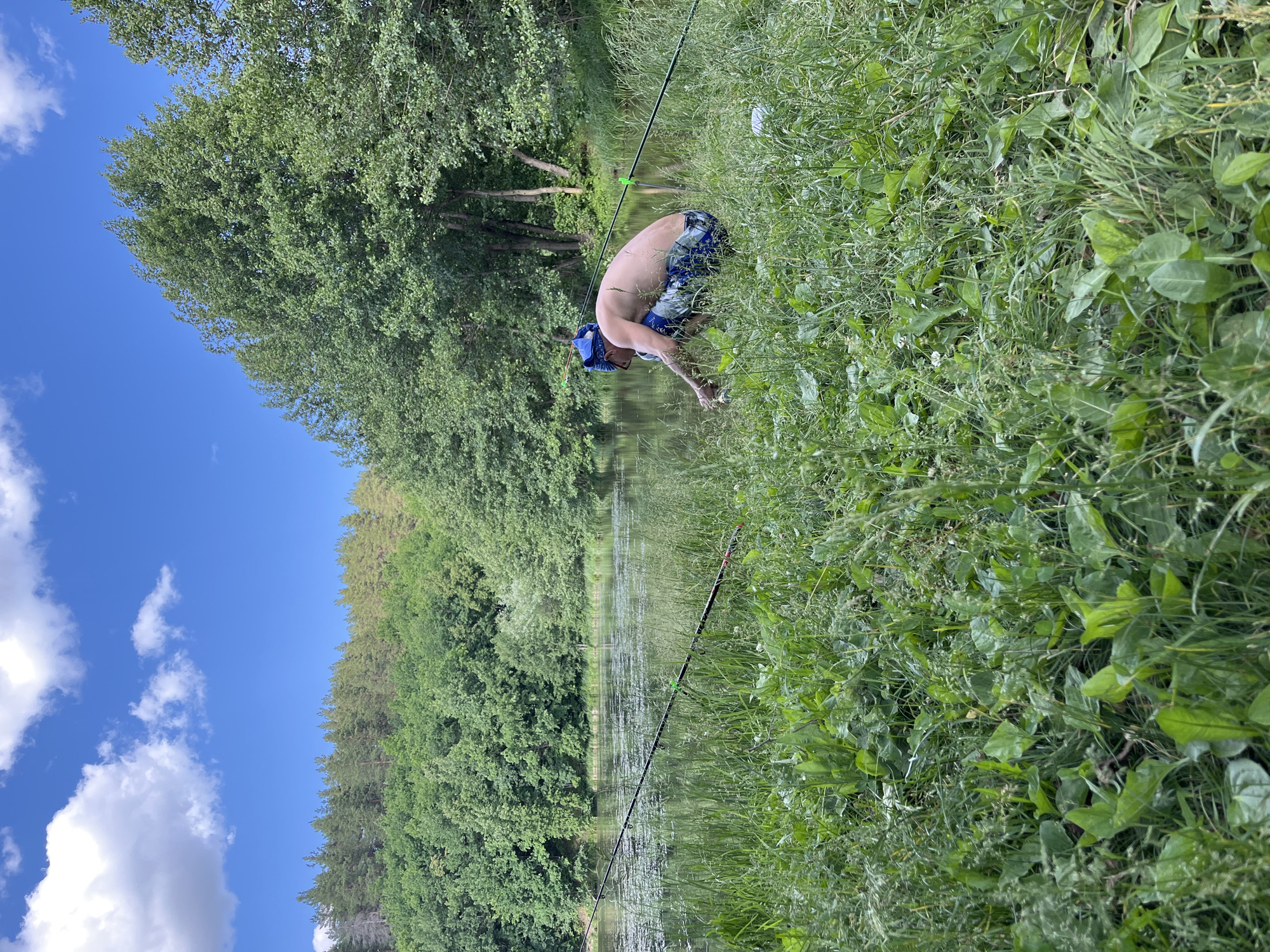 A shirtless man wearing a blue bandana and patterned shorts is crouched on the grassy bank of a lake, holding a fish. Two fishing rods are set up in the foreground, with their tips resting in green holders planted in the ground. The lake is surrounded by lush green trees, and the sky above is blue with scattered white clouds. The scene appears to be a natural, outdoor setting.