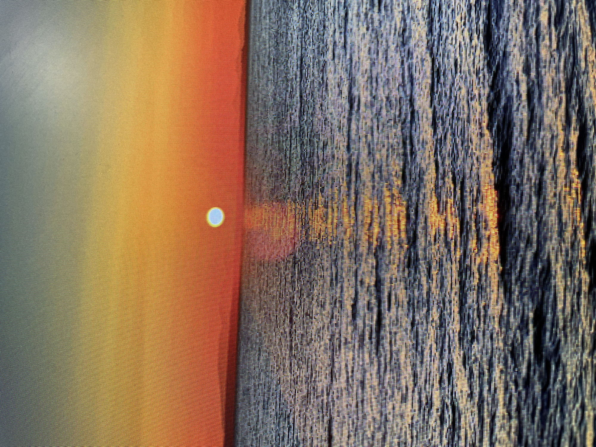 The image appears to be a close-up photograph of a screen displaying a sunset over water. The left side of the frame shows a gradient of colors transitioning from gray to yellow to orange, with a bright, circular light source representing the sun. The right side depicts water, with vertical striations and reflections suggesting waves or ripples catching the light. The overall texture is pixelated due to the close-up view of the screen. There is no evidence to suggest that this image is of the city of Zlatograd, Bulgaria.